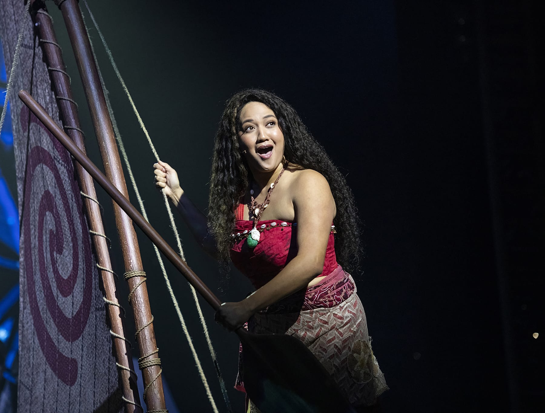 A performer in a red and brown outfit sings on stage, holding a rope against a backdrop resembling a sail, evoking the adventurous spirit of Moana during the Disney Treasure Cruise Ship's captivating stage show.