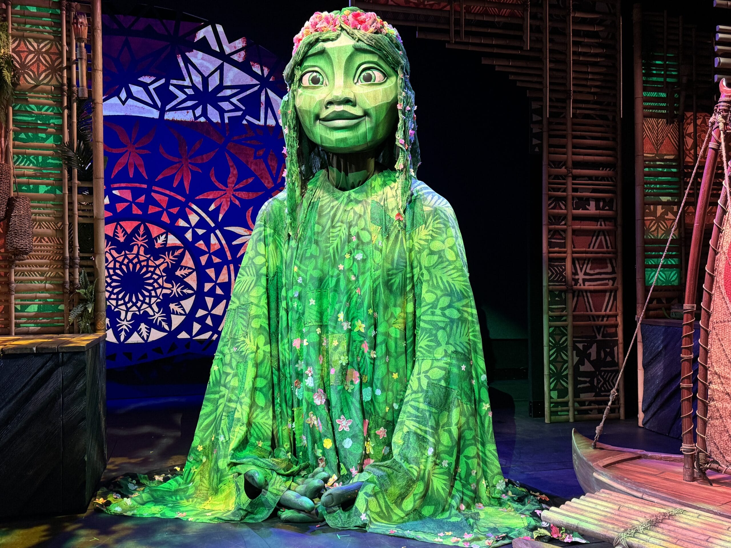A large green puppet with floral patterns and a headdress sits on a stage, evoking the magic of a Moana-themed stage show. Intricate designs in the background add to the Disney Treasure Cruise experience.