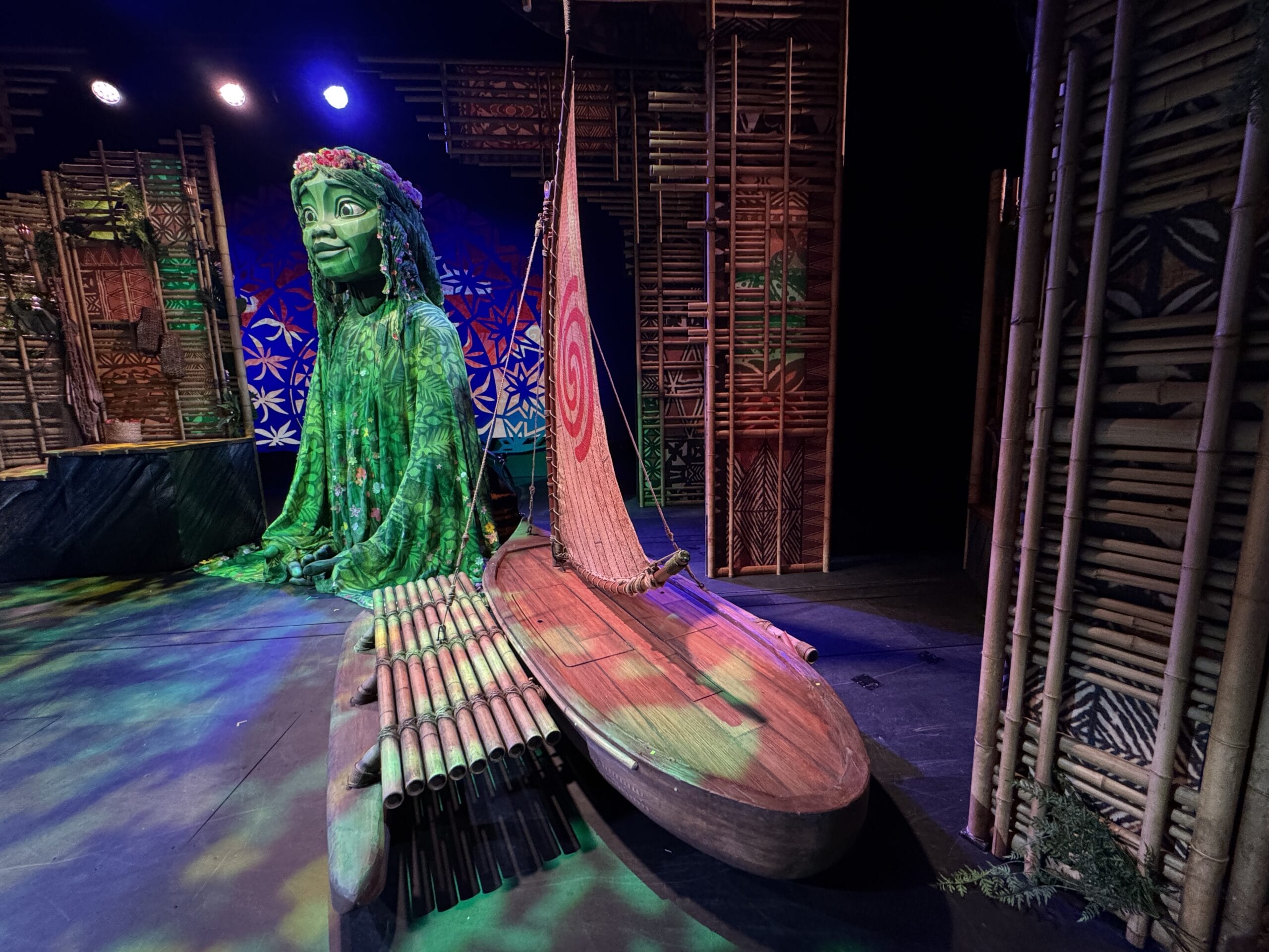 The stage set, reminiscent of a Moana scene, features a large green figure resembling a seated goddess with long hair, a wooden boat sporting a spiral sail, and bamboo decor, all brilliantly illuminated by stage lights. This enchanting setup beckons you into the magical world of Disney's Treasure Cruise.