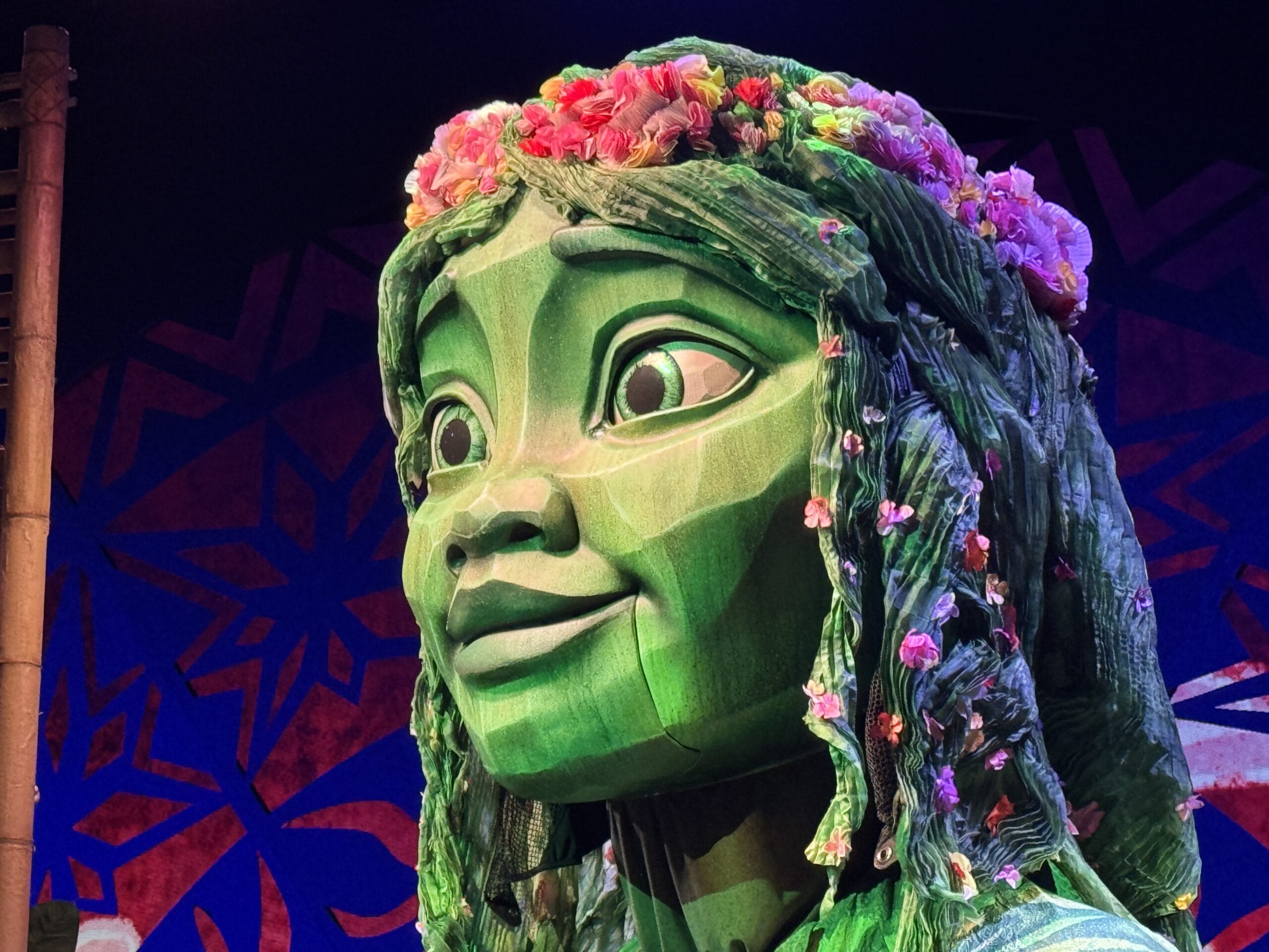 A large green puppet adorned with a floral crown stands majestically against a geometric blue and red backdrop, evoking the enchanting atmosphere of a Moana-inspired stage show on the Disney Treasure.