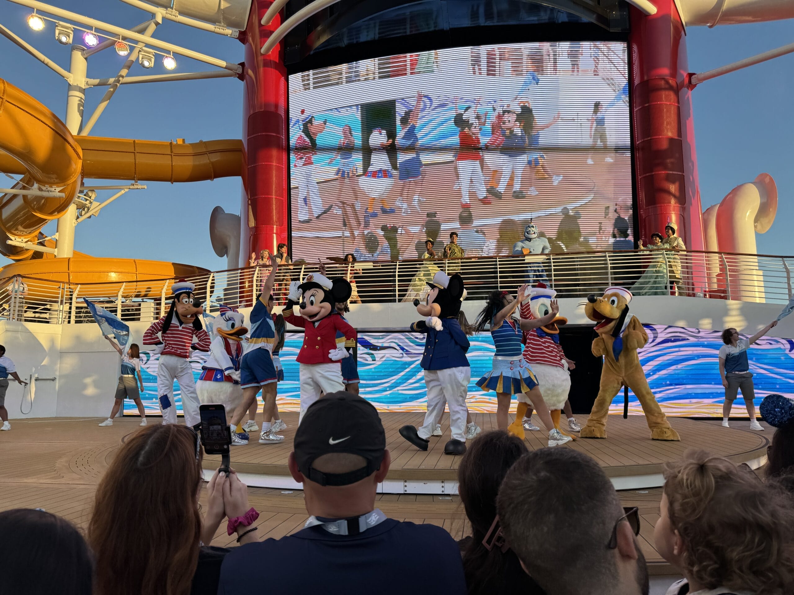 Characters in costumes perform on the cruise ship deck stage, enchanting the audience with Disney treasure wonders. A large screen displays animated characters in the background, setting sail into a magical voyage of entertainment.