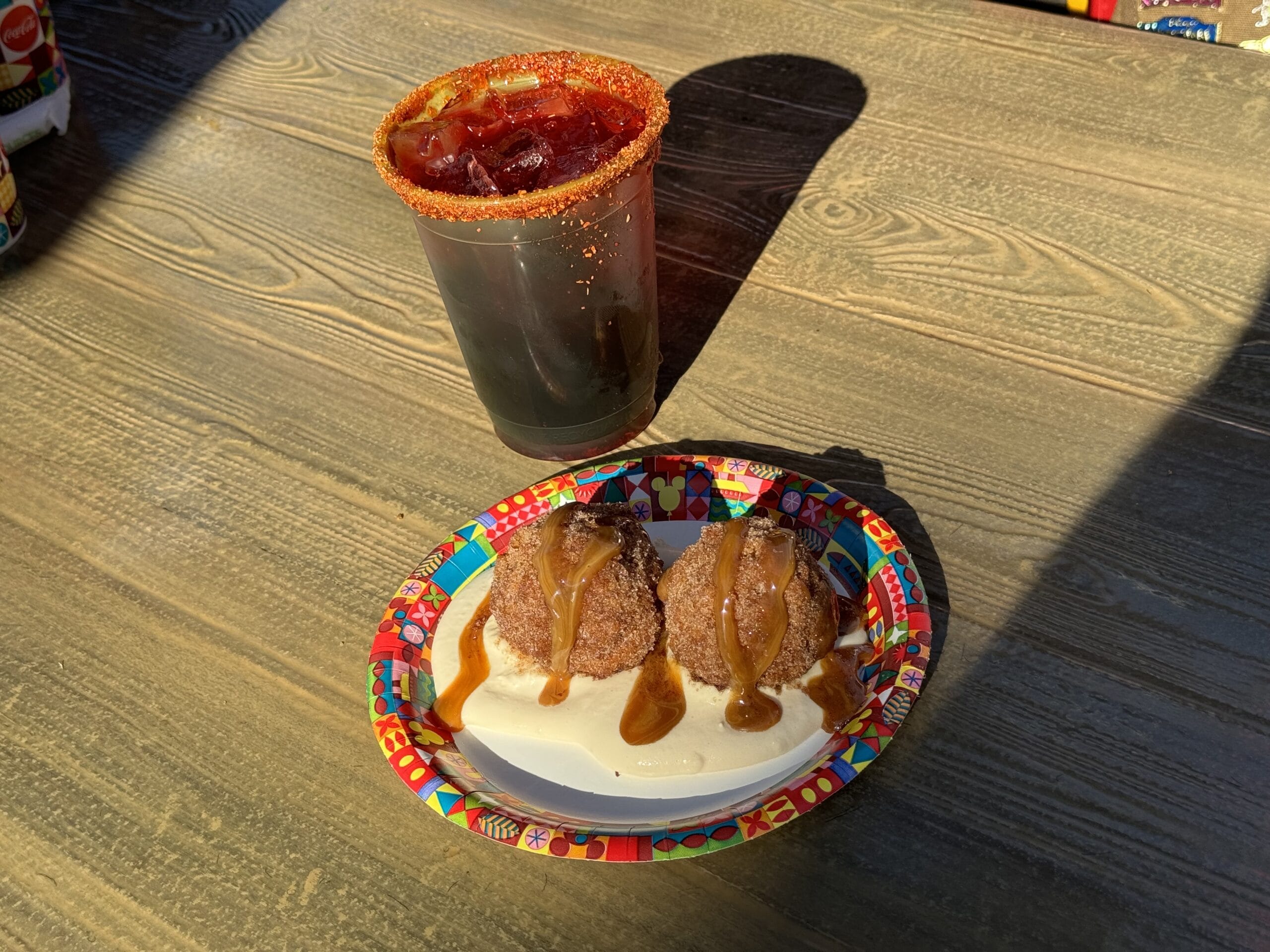 A cup with a red, chili-rimmed drink from Cocina Cucamonga sits next to a colorful plate containing two brown desserts topped with caramel sauce, all resting on a wooden surface.
