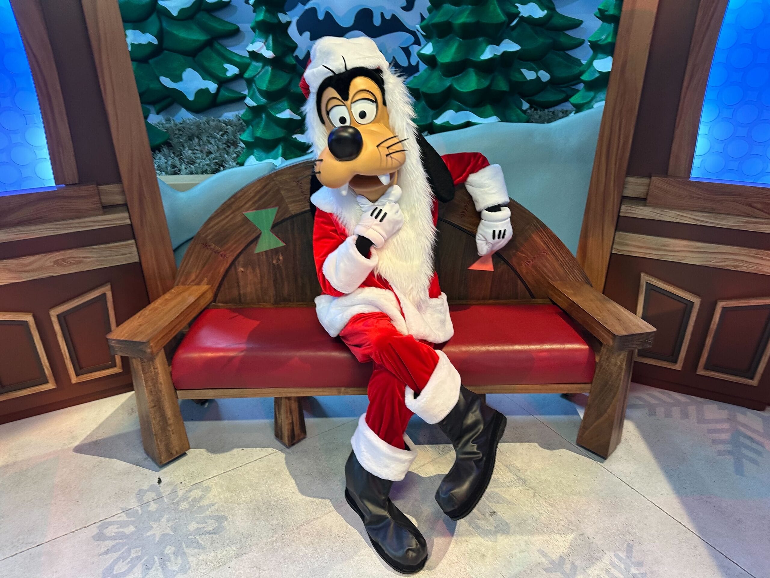 A costumed character dressed as Santa Goofy sits on a wooden bench in front of a snowy backdrop with trees, capturing the whimsical charm of Fantasyland Theatre at Disneyland Park.