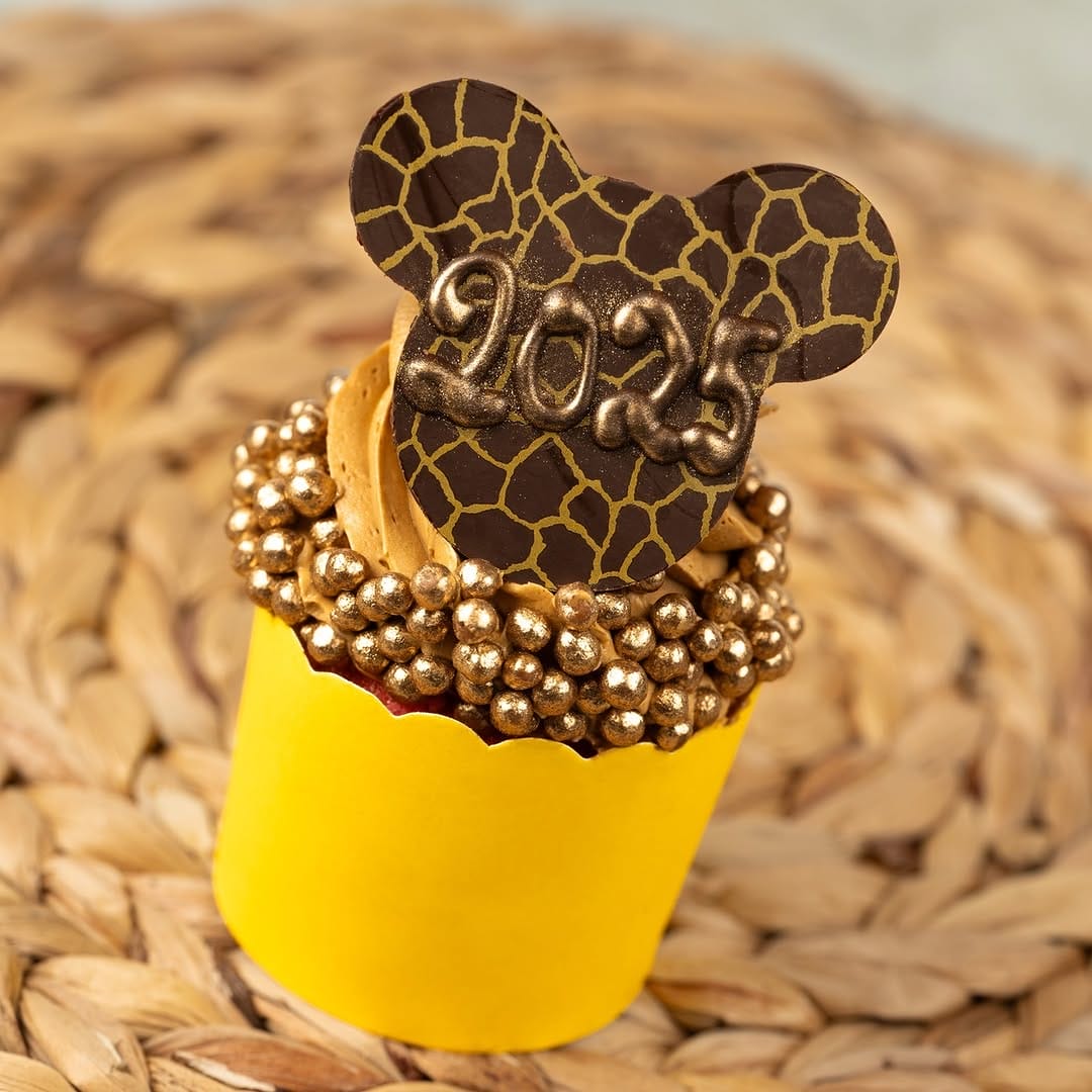 Discover a delightful cupcake featuring gold beads and a chocolate topper resembling mouse ears, adorned with "2023" text. This exclusive Disney Parks treat comes in a yellow wrapper and rests on a woven surface, perfect for celebrating the upcoming New Year's Eve 2025.