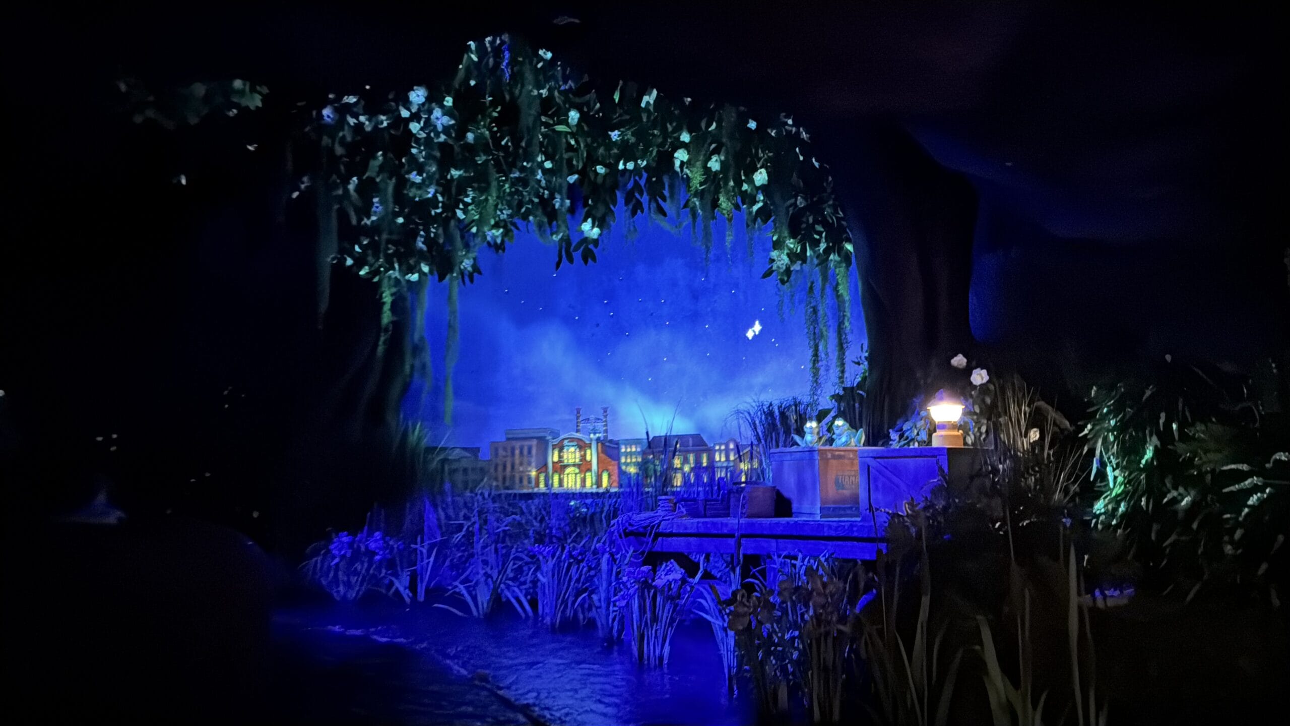 A dimly lit diorama captures a small town under a starry night sky, reminiscent of the Magic Kingdom. Hanging vines frame the scene, while illuminated buildings in the background add an otherworldly glow to this enchanting display.