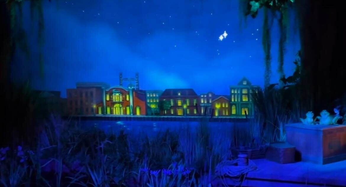 Night scene of a lit-up waterfront town with glowing buildings against a starry sky. Water reflections and silhouetted vegetation enhance the scene.