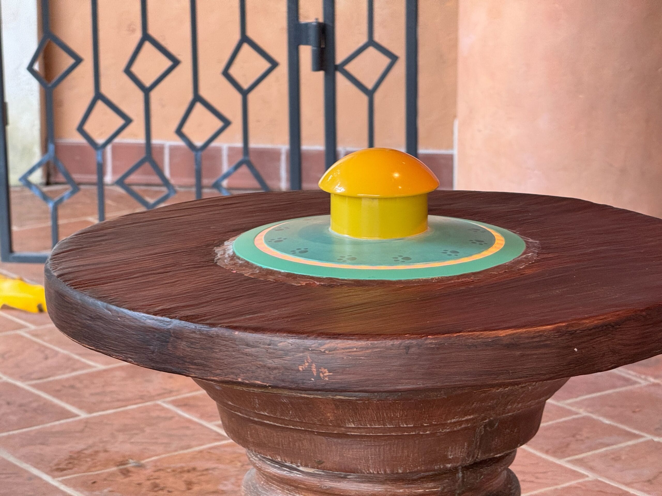 Wooden table with a small, round, yellow plastic object on top, set against a tiled floor and metal fence background.