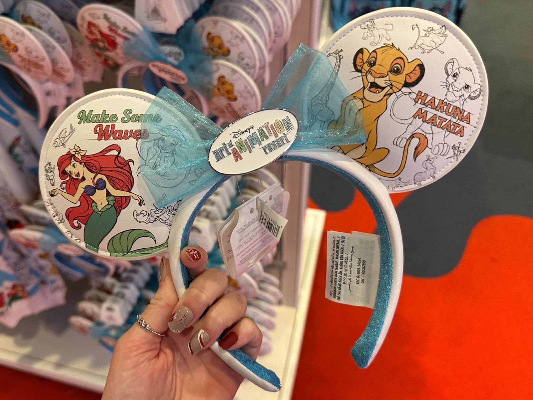 A person holds a pair of themed mouse ears with "Hakuna Matata" and "Make Some Waves" designs.