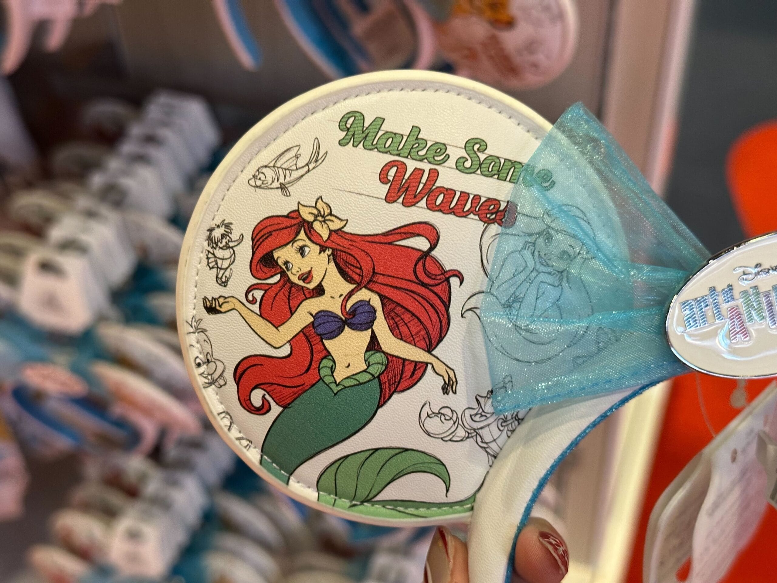 A circular badge featuring a red-haired mermaid with the text "Make Some Waves." The background includes small sea creatures and a blue bow on the side.