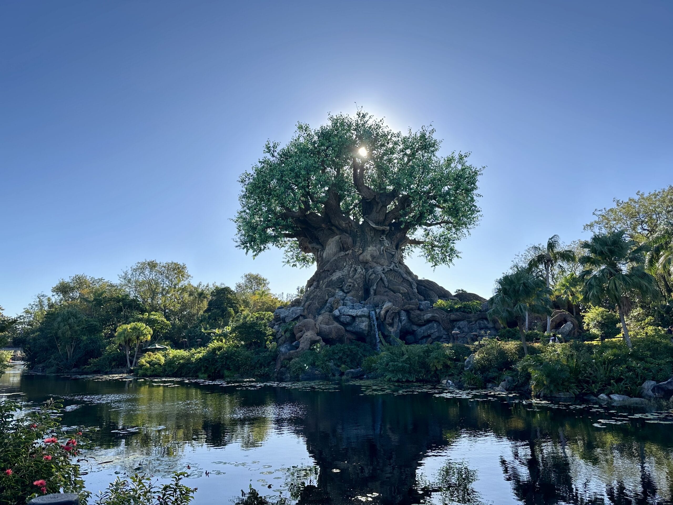 A large sculpted tree with intricate carvings stands by a reflective body of water, surrounded by lush greenery under a clear blue sky.
