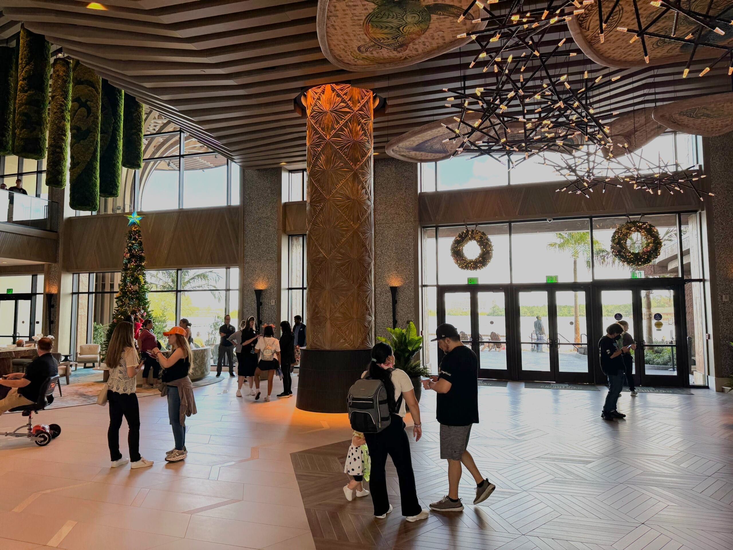 The DVC Island Tower hotel lobby buzzes with people walking and talking, beautifully adorned for the season with a grand Christmas tree and festive wreaths. A large patterned column stands tall, complementing the modern lighting that casts a warm glow over this holiday oasis.
