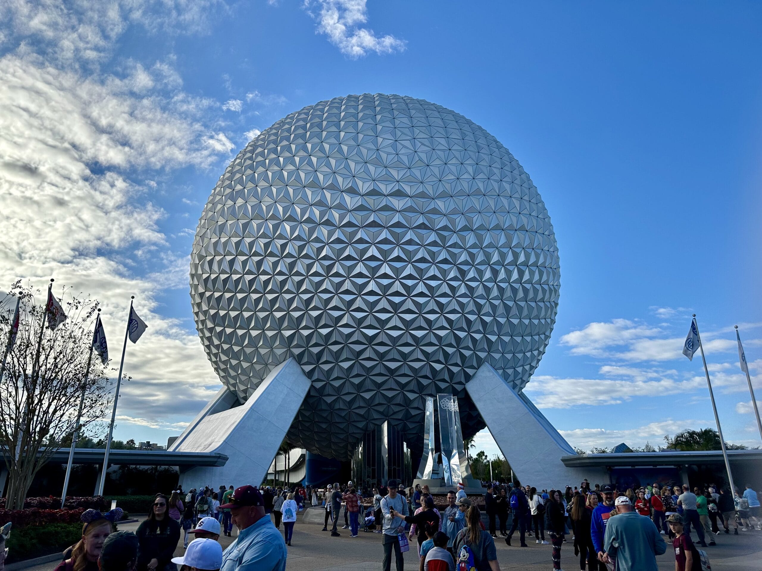 A large geodesic sphere at Epcot, surrounded by people and flags, under a partly cloudy sky.