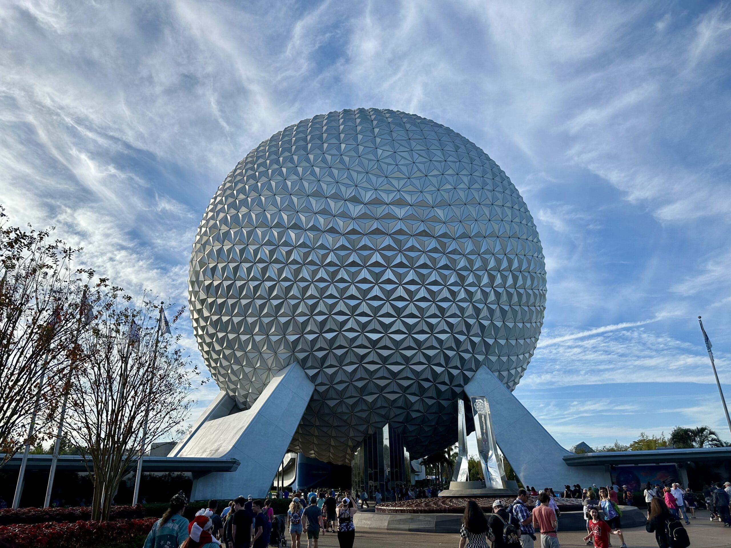 Spaceship Earth, a large geodesic sphere, stands prominently at Epcot, with a crowd of visitors below and a partly cloudy sky above.