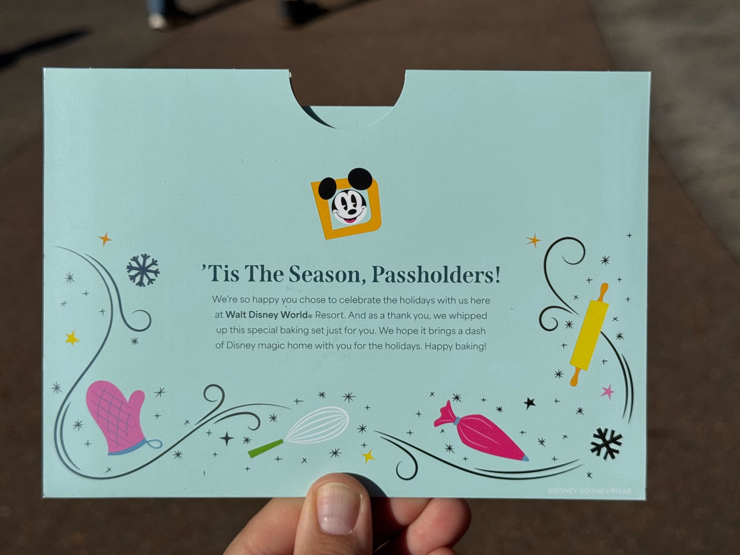 A hand holds a Disney envelope adorned with holiday graphics and a special message for EPCOT passholders about receiving a free recipe card as a unique baking gift.
