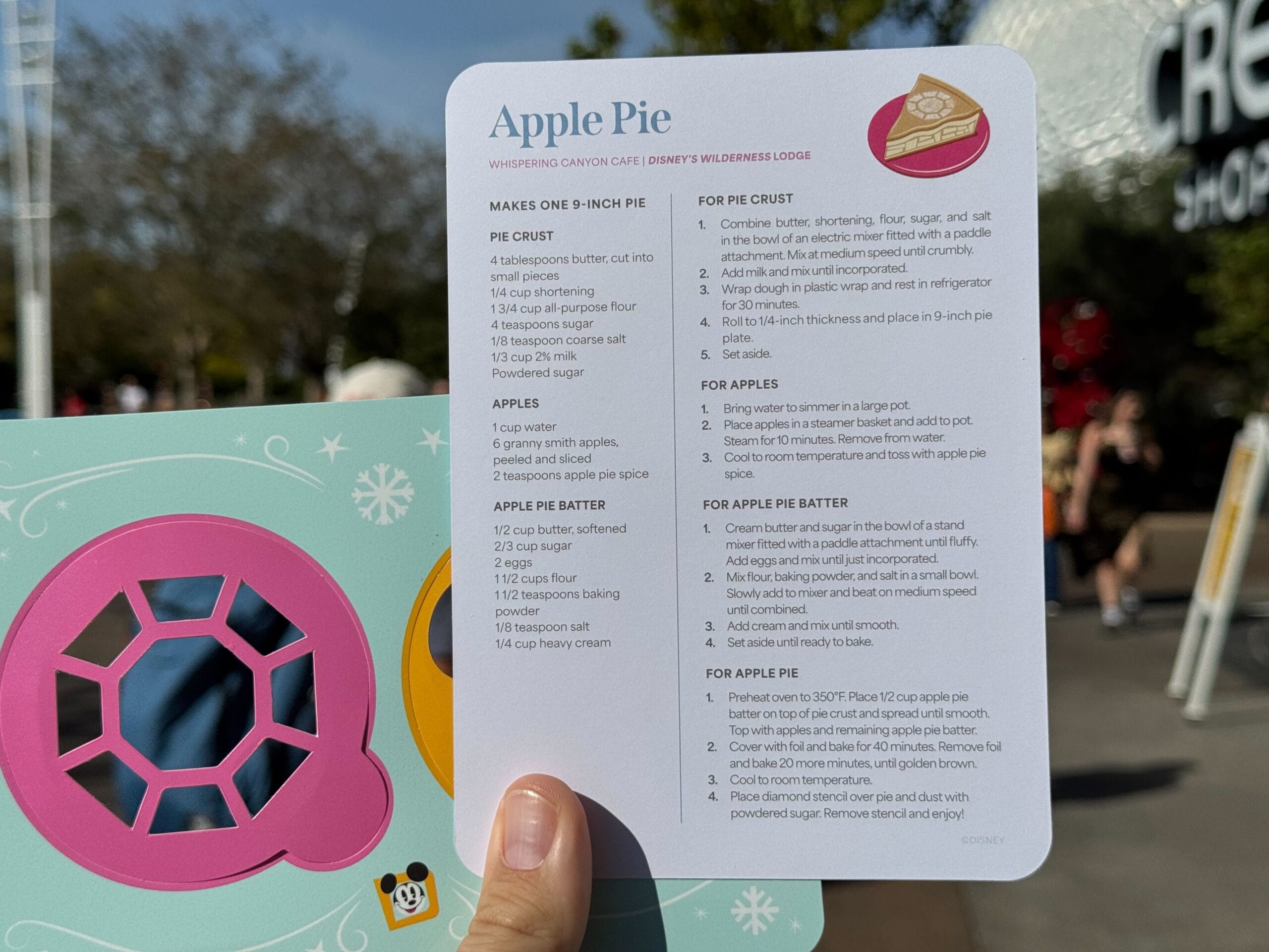 An Annual Passholder is holding an Apple Pie recipe card outdoors at EPCOT, with another colorful free recipe card partially visible in the background.