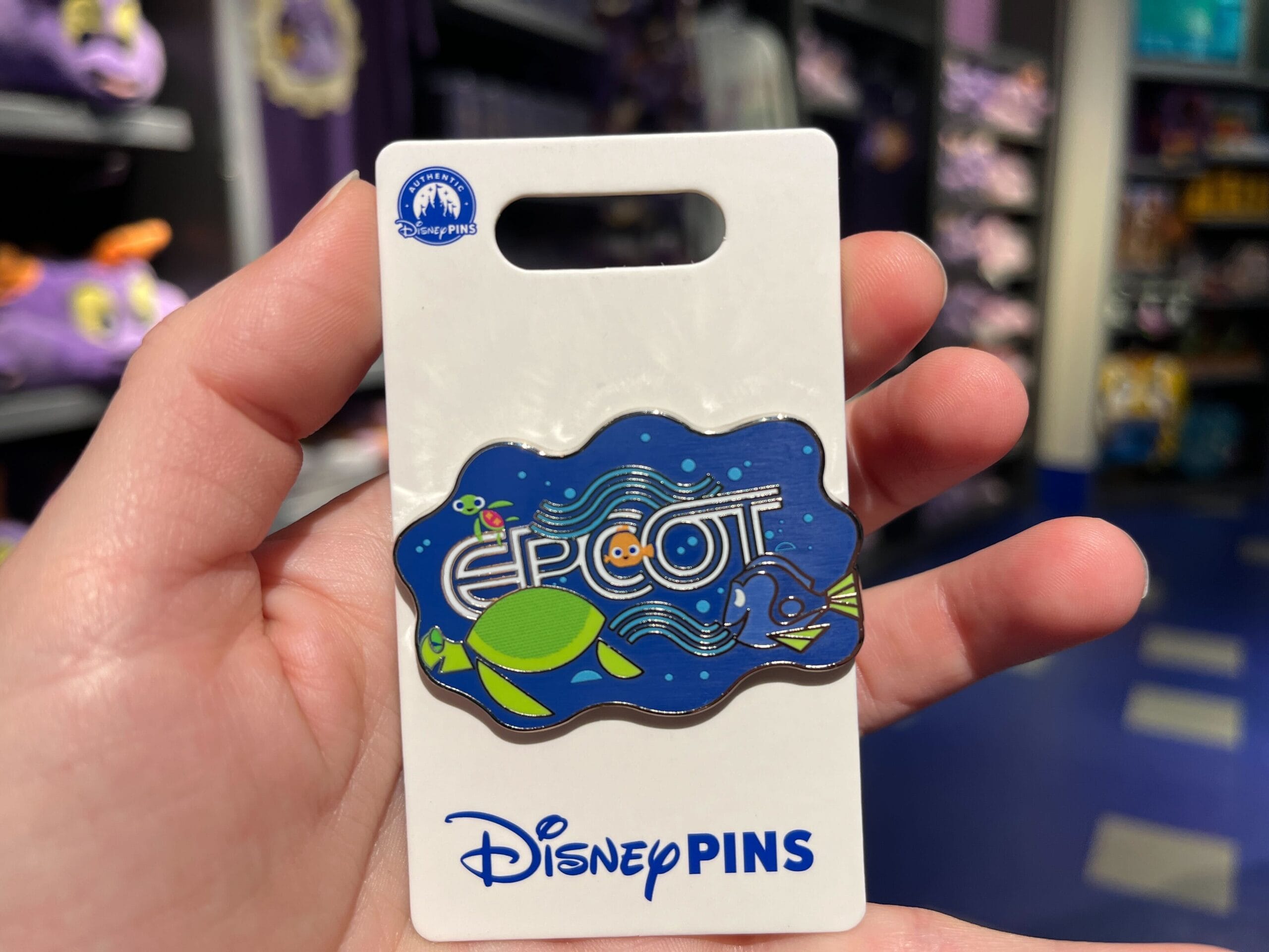 A hand holds a Disney pin featuring a blue "Epcot" design with sea turtle and fish illustrations on a display card labeled "Disney Pins.