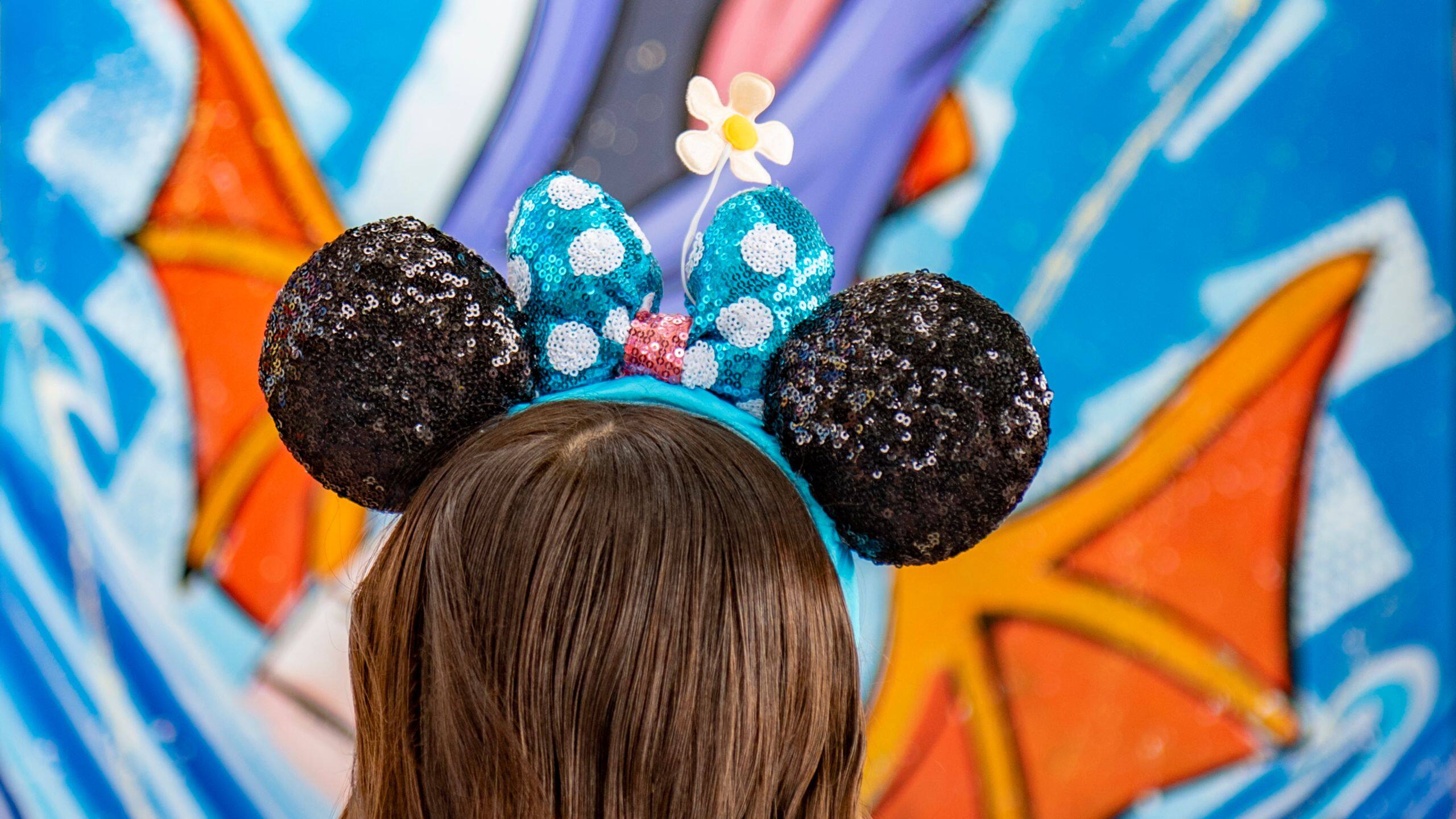 A child wearing sequined Mickey Mouse ears headband with a flower stands joyfully against a vibrant, festival-inspired abstract wall art.