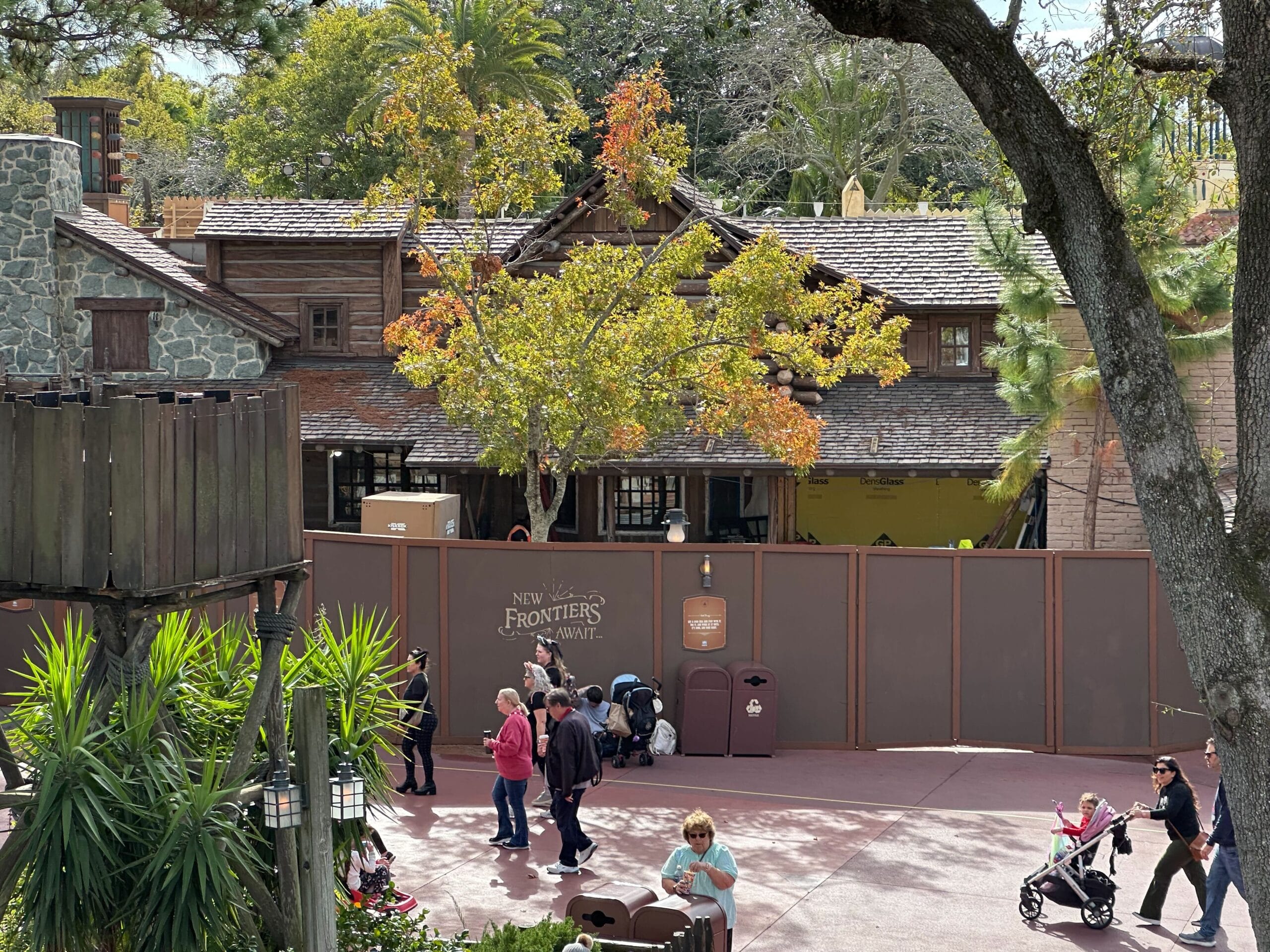 Construction walls labeled "New Frontiers Await" with people walking by. A rustic building and trees are visible behind the walls.