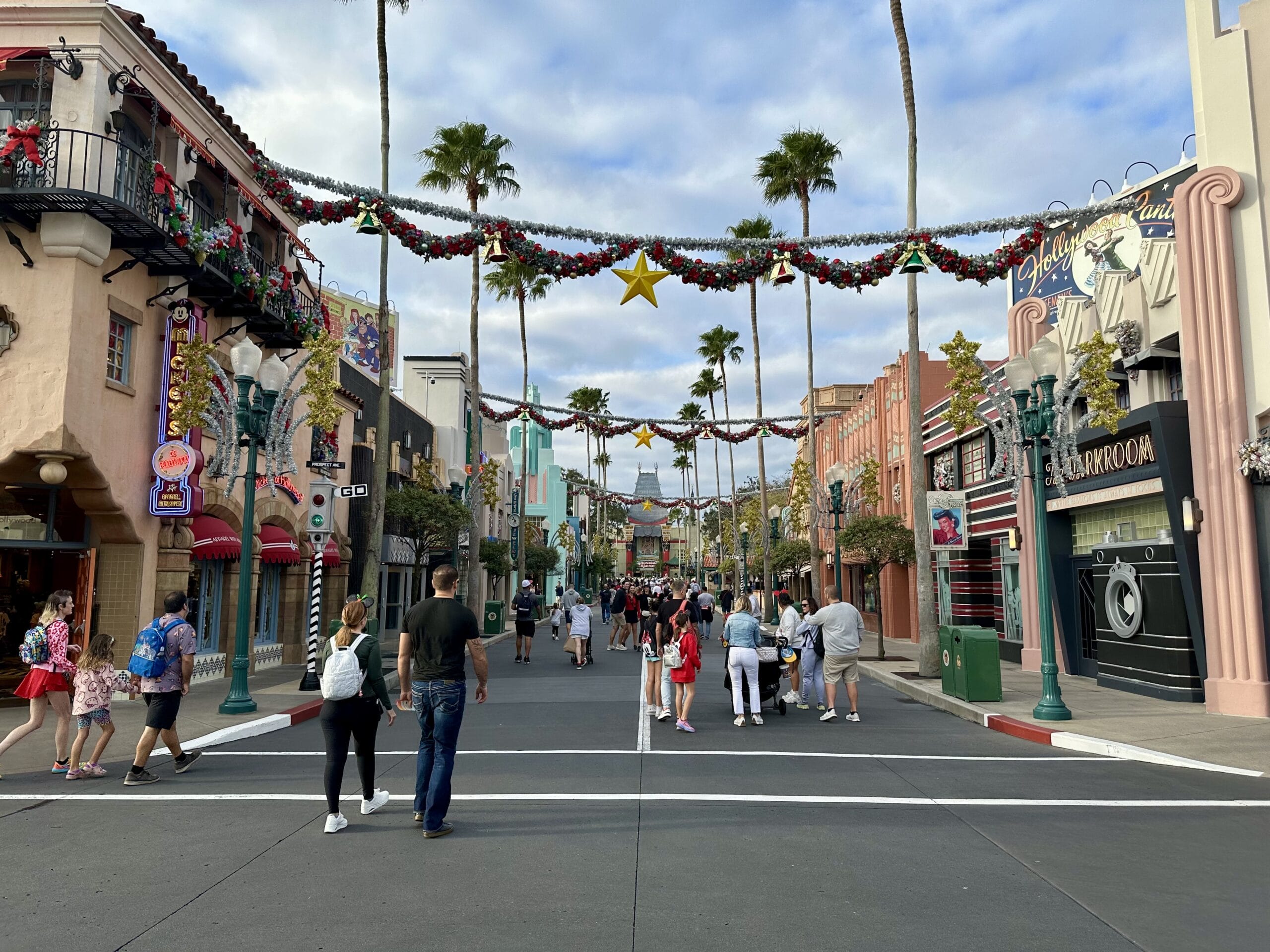 A street scene with people strolling among decorations, including garlands and stars, flanked by themed shops and palm trees under a partly cloudy sky, reminiscent of the vibrant atmosphere found at Disney's Hollywood Studios.
