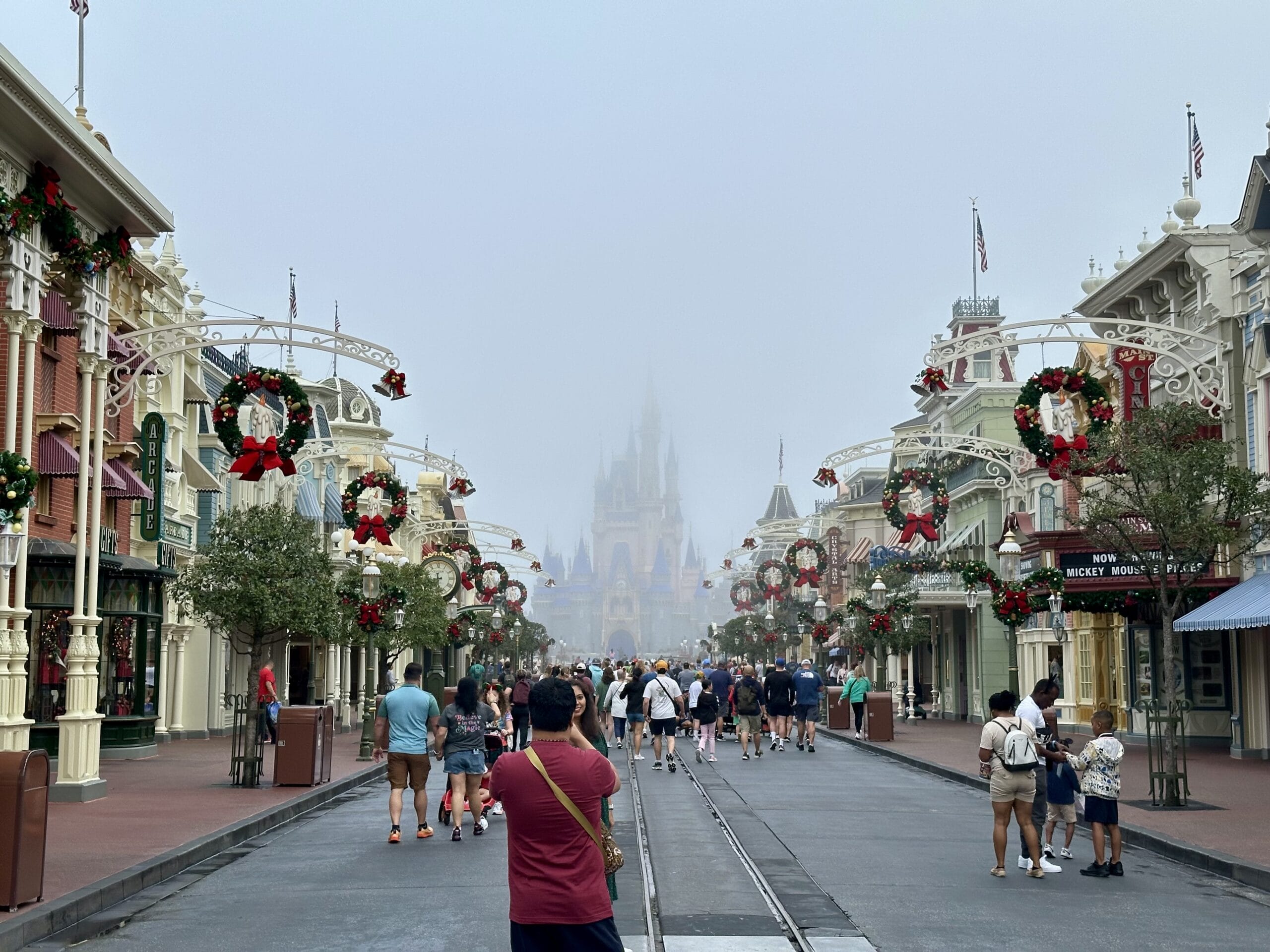 People walk down a festive, foggy street lined with decorated buildings, leading to a distant castle.