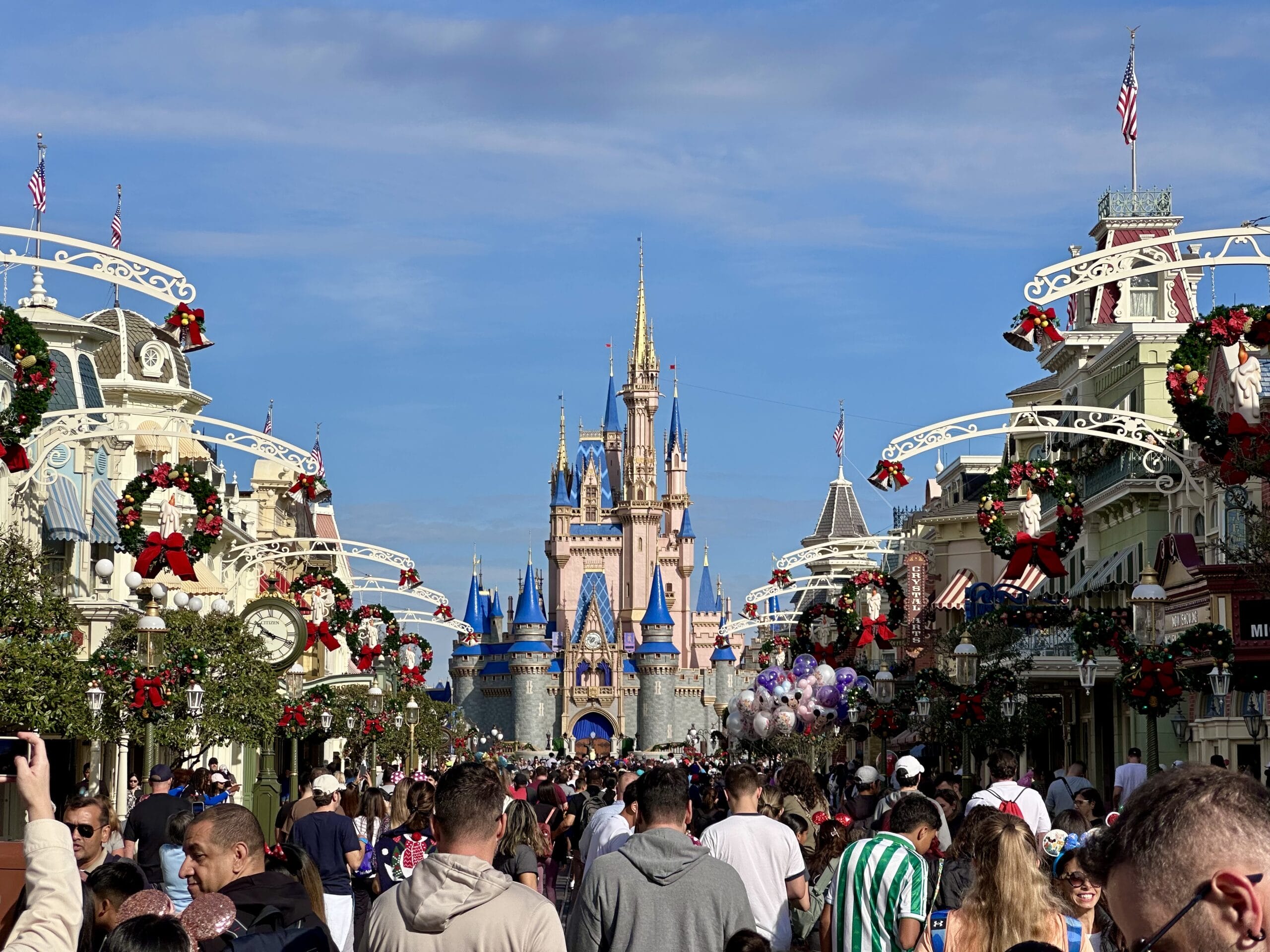 A crowded Main Street at a theme park features festive decorations and a castle in the background under a clear blue sky.