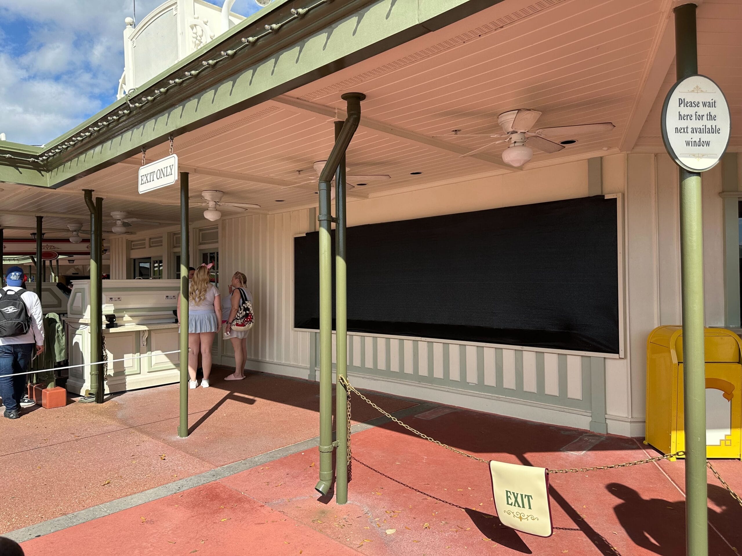 People stand near a closed service window at an outdoor building with a rope and "exit only" signs.