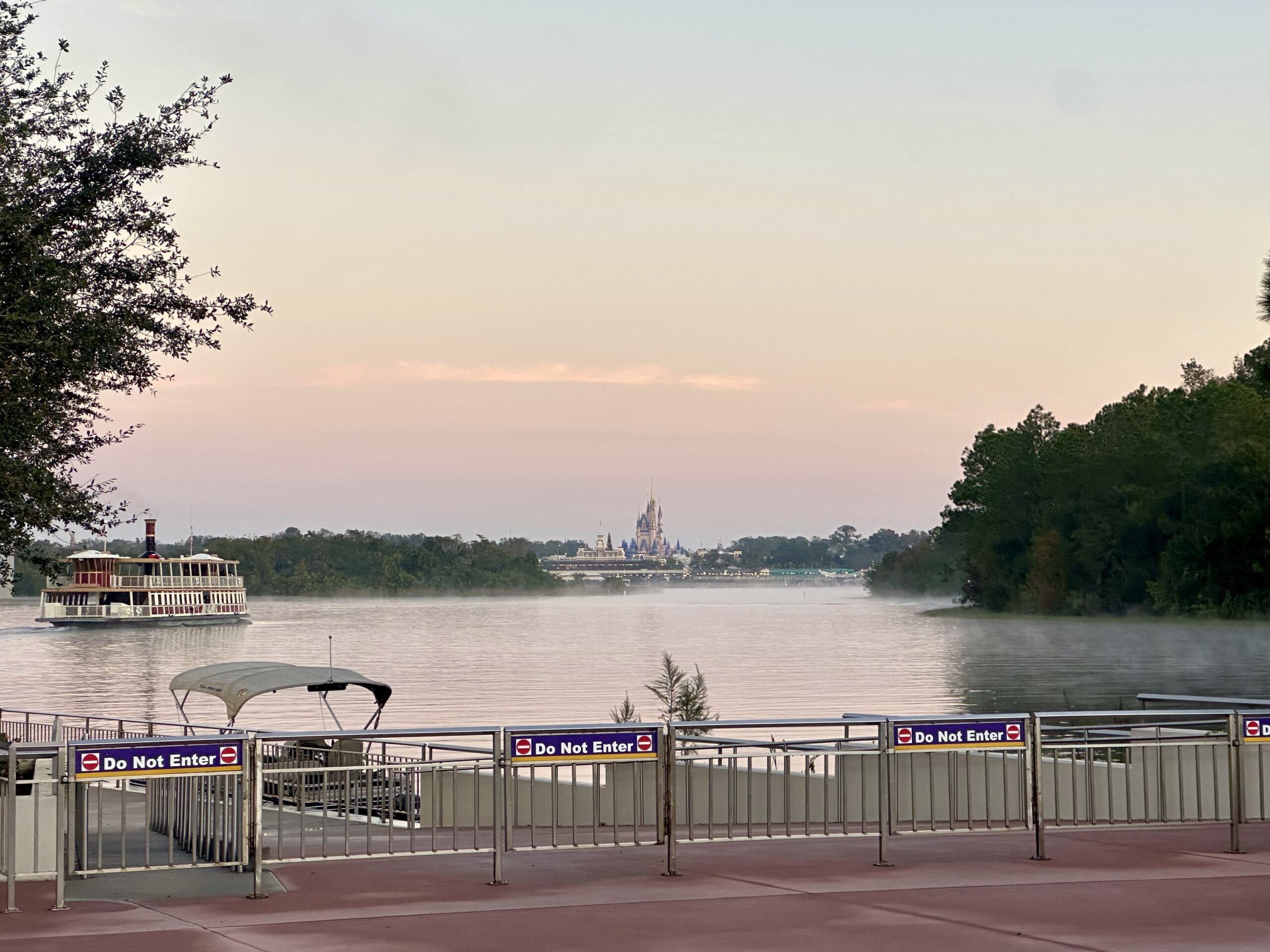 A river scene at dawn with a docked boat and a ferry on the water. A distant castle is visible under a pastel sky. Signs on the dock read "Do Not Enter.