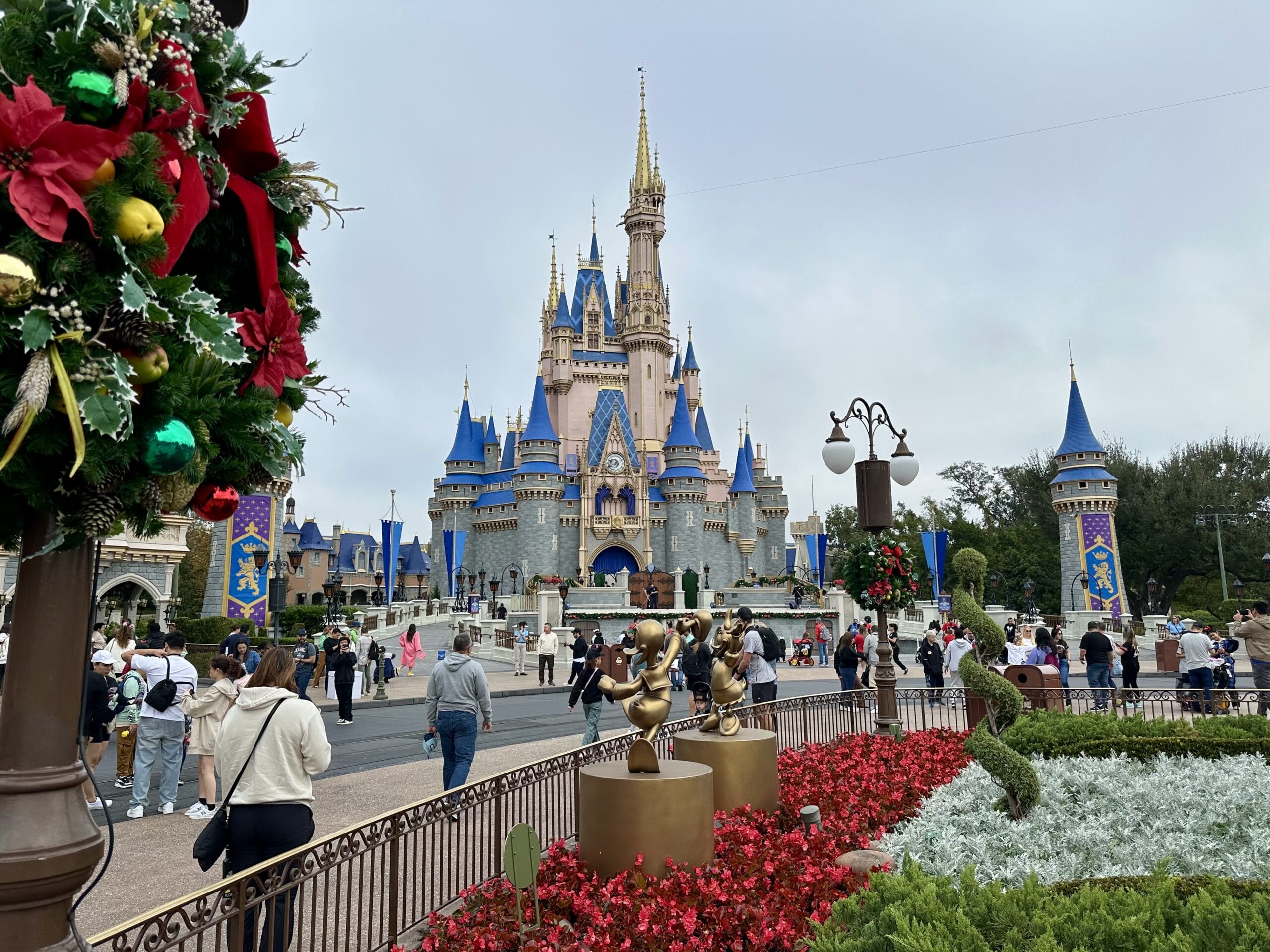 Visitors explore a theme park with a castle in the background, surrounded by flowers, a festive wreath, and statues of cartoon characters.