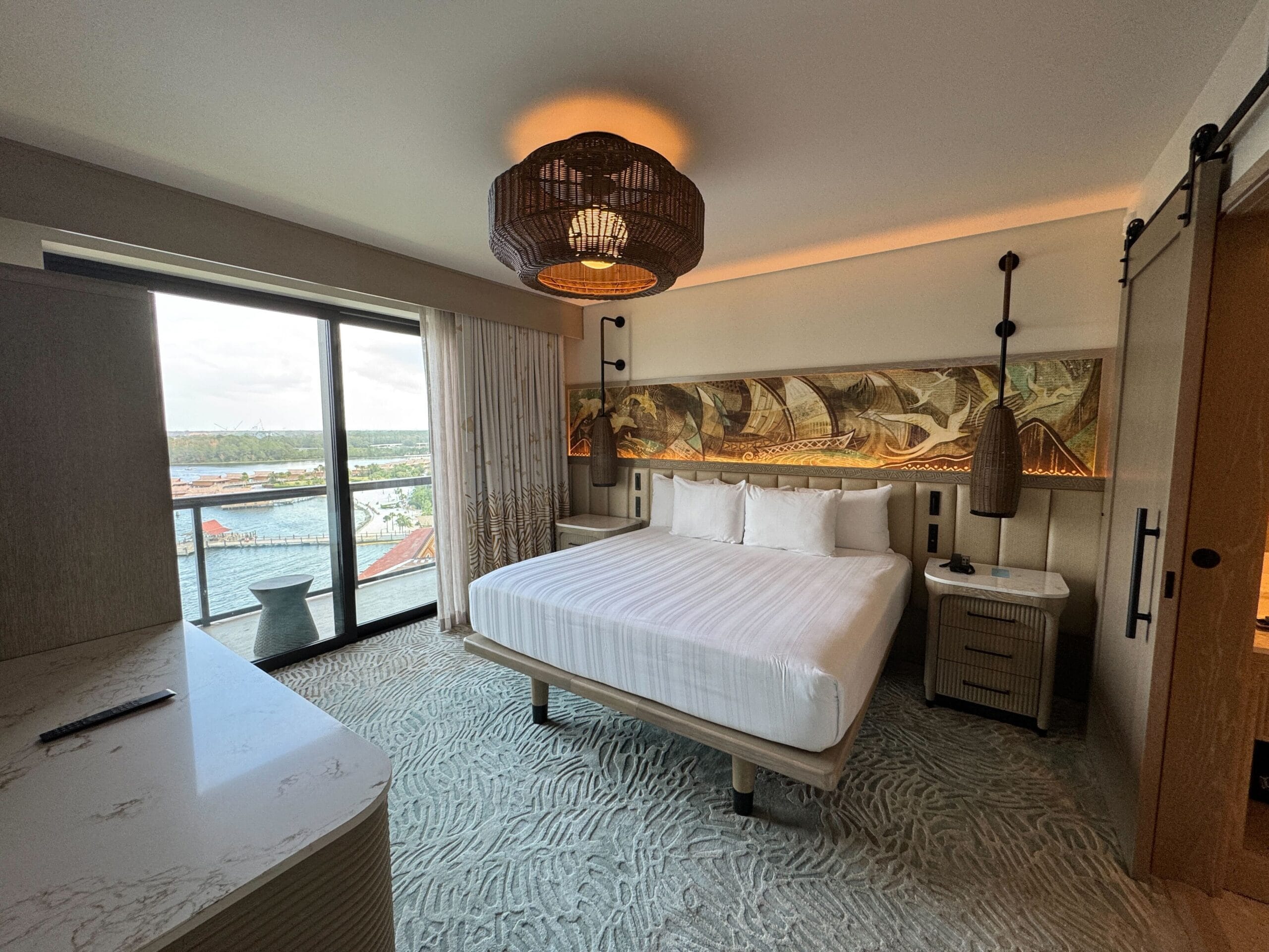 Experience the charm of our Polynesian-inspired hotel room, featuring a king-size bed, decorative artwork, and a wicker light fixture. Bask in the serenity offered by a large window revealing an ocean view.