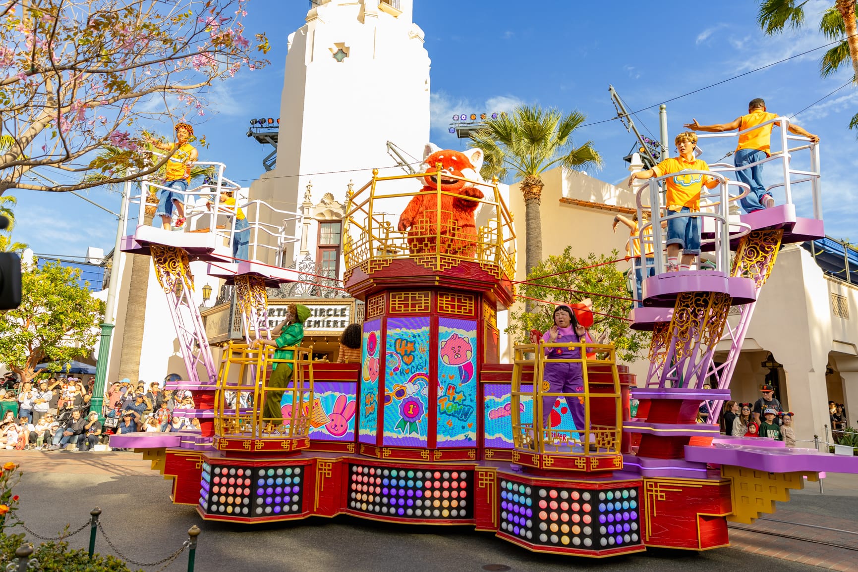Colorful parade float featuring performers and a character in costume, surrounded by a crowd under a clear blue sky.