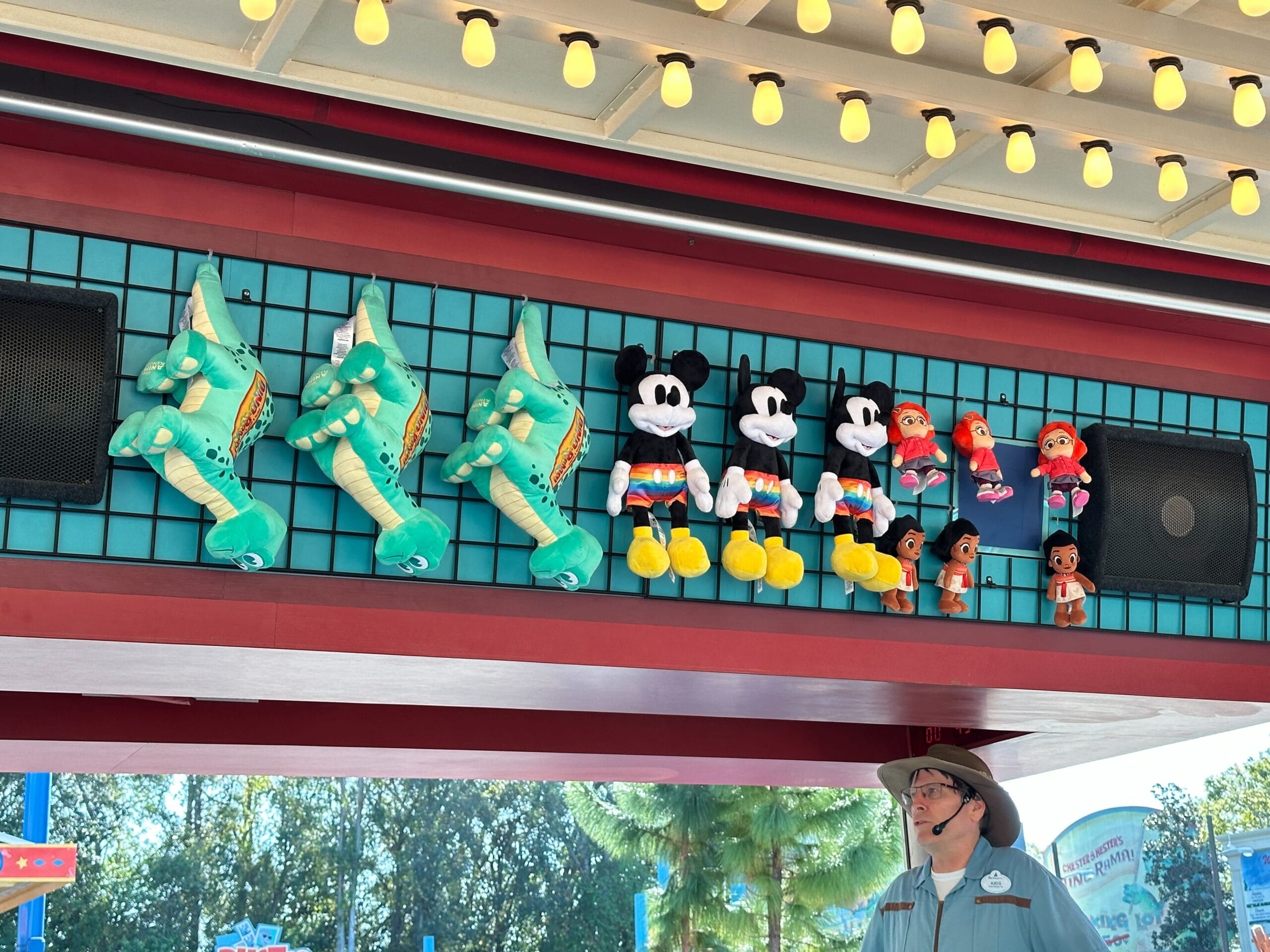 A DinoLand Carnival Games display showcases stuffed toy prizes like dinosaurs and cartoon mice, alongside Wishables in red outfits. A staff member stands below, overseeing the lively scene.