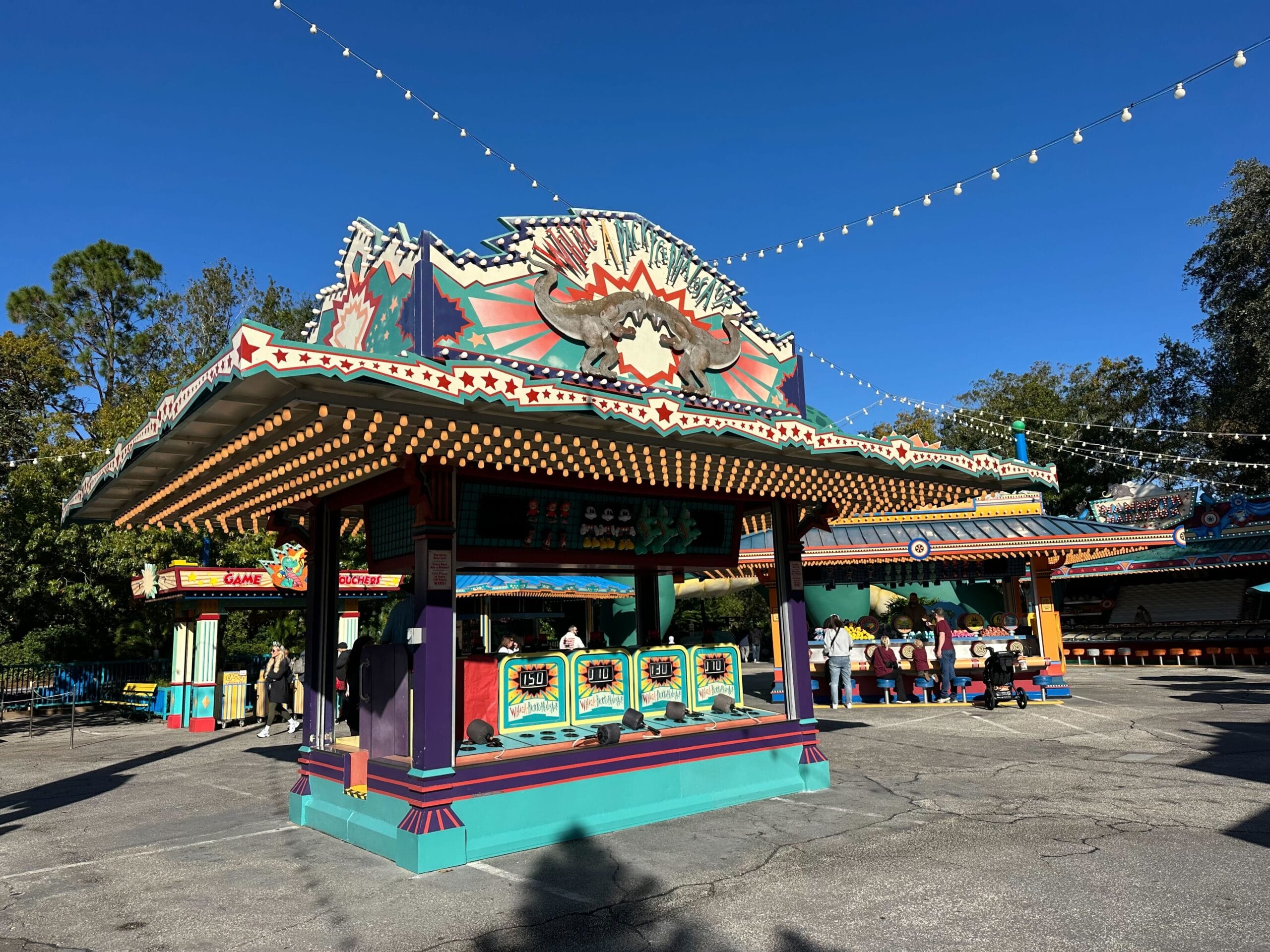 The DinoLand Carnival Games booth bursts with vibrant colors and twinkling lights under a clear blue sky, as excited visitors gather nearby, eager to win Wishables and other delightful prizes.