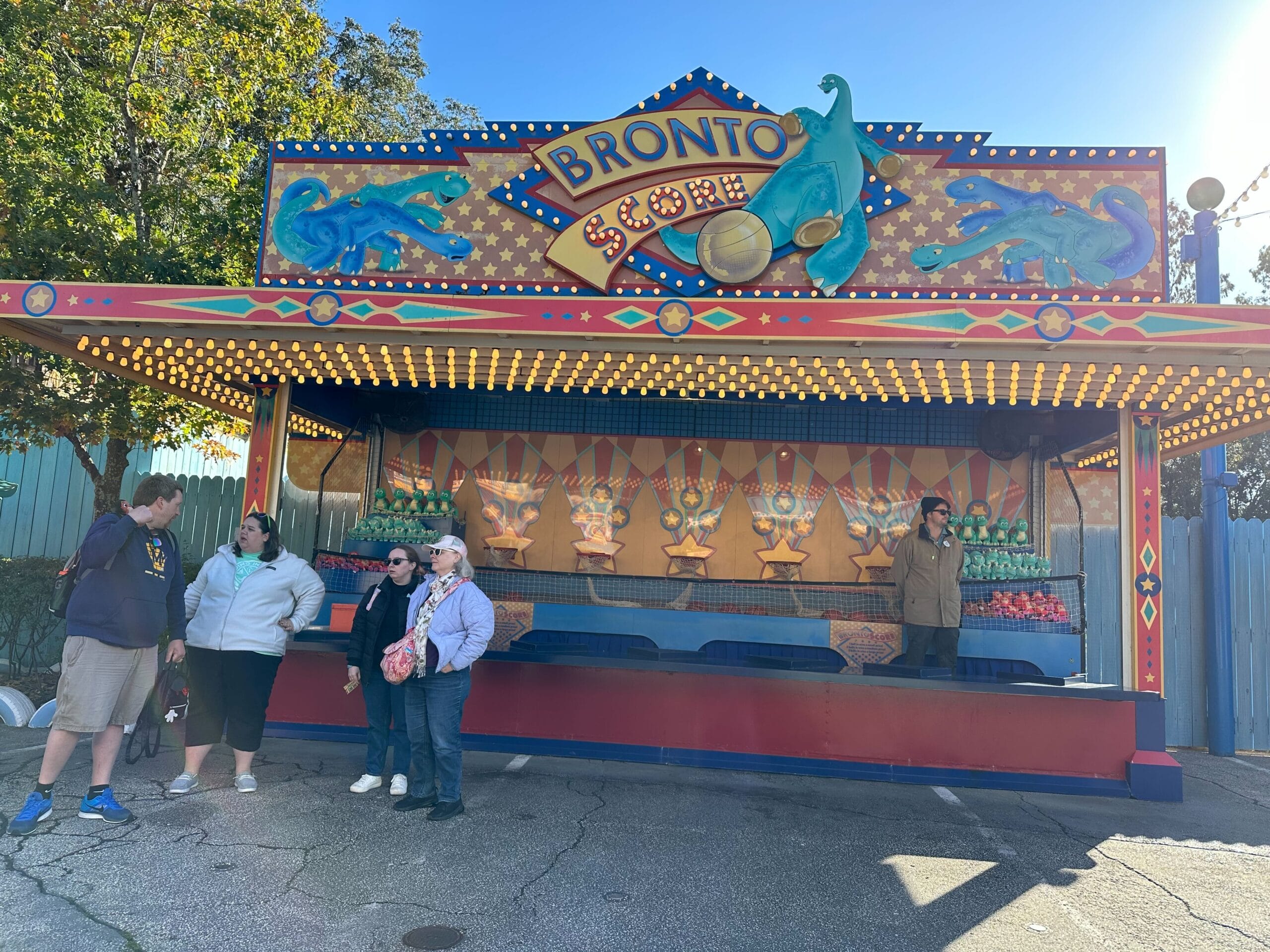 People stand in front of a carnival game booth called "Bronto Score," set against the clear blue sky, with dinosaur art that's reminiscent of Dinoland's whimsical charm.