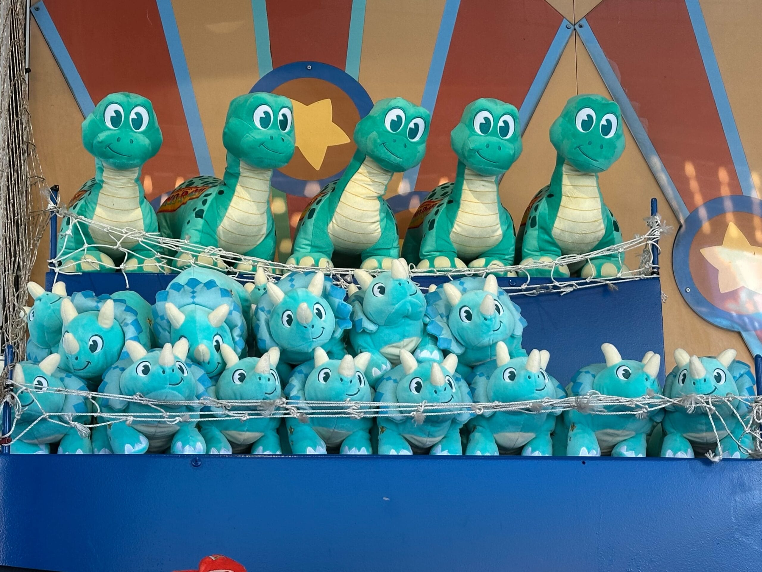 Two shelves display plush Wishables dinosaur toys, with turtles on the top shelf and triceratops on the bottom. The background resembles DinoLand Carnival Games, adorned with a colorful, starry design.