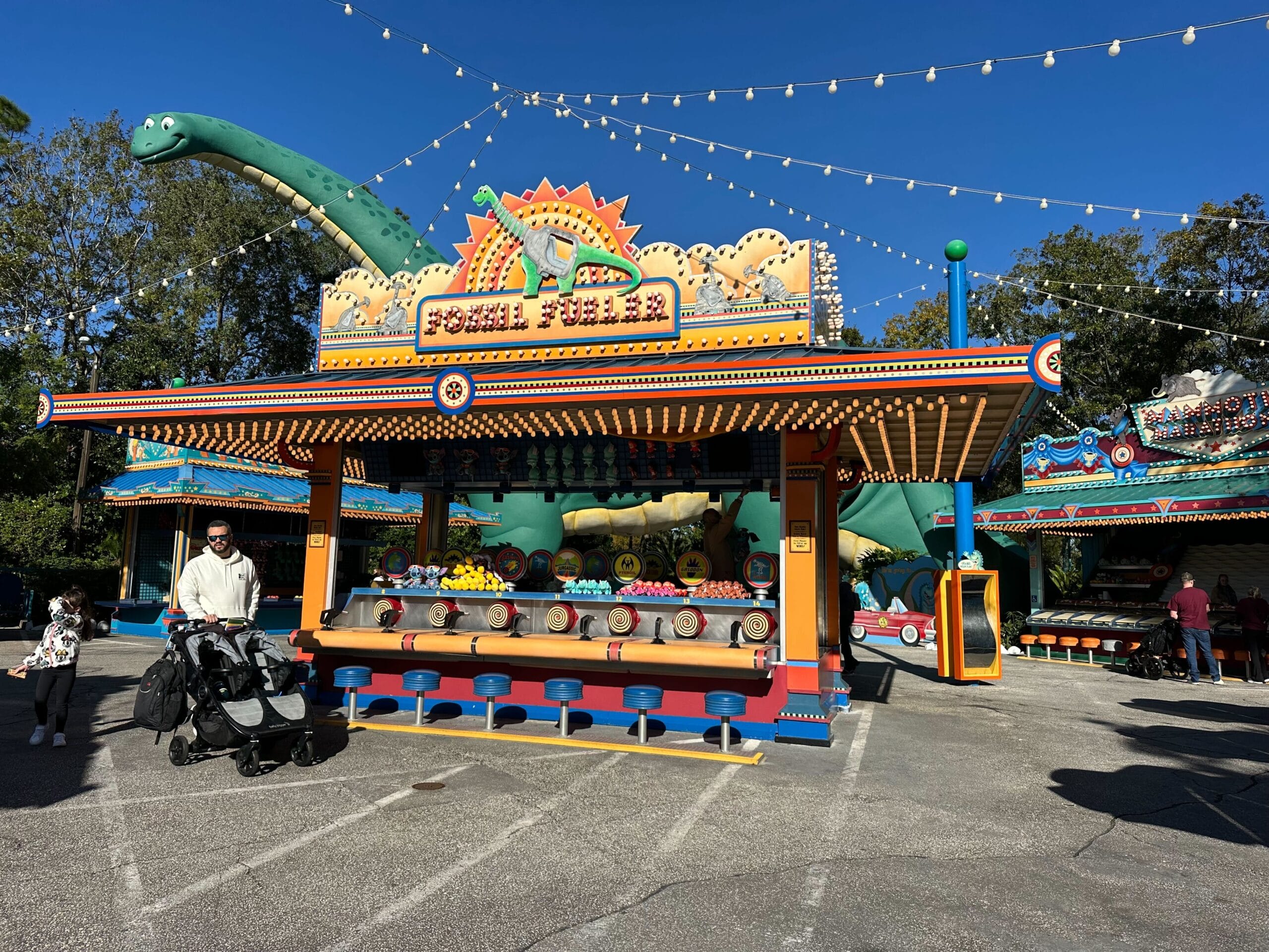 A colorful carnival game booth at DinoLand in the amusement park, featuring a dinosaur theme. A person with a stroller stands nearby under sparkling string lights, filled with the cheerful buzz of Carnival Games.