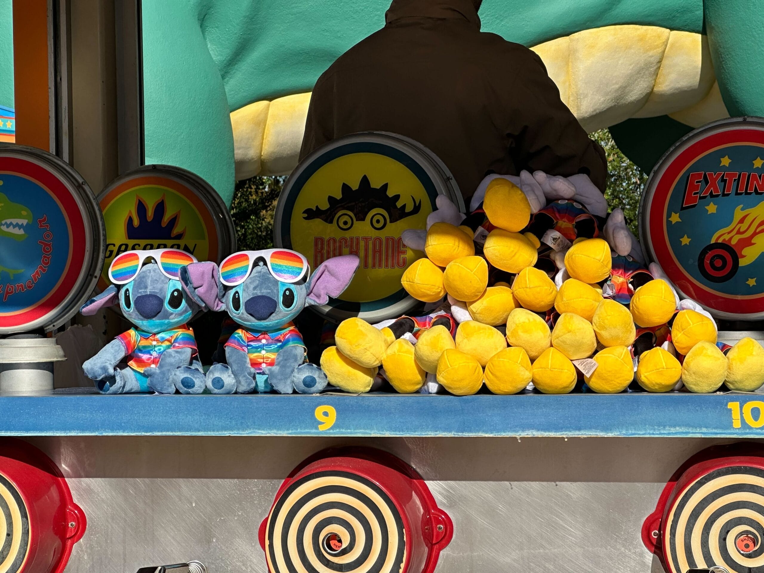 Stuffed toy prizes, including two plush creatures wearing sunglasses and colorful shirts, are displayed on a carnival game shelf beneath vibrant, playful DinoLand signage.