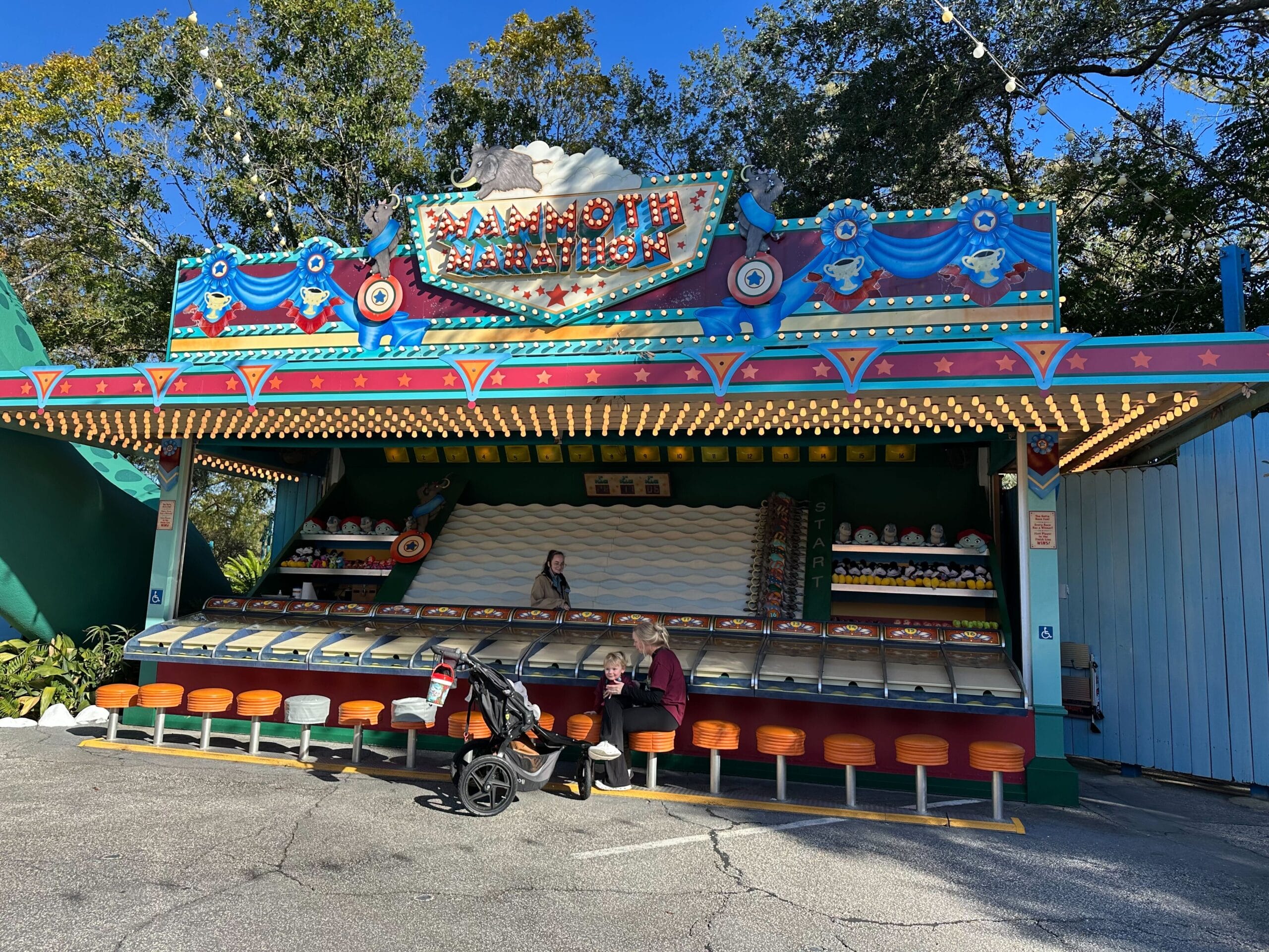 An empty carnival game booth, labeled "Mammoth Marathon," displays vibrant animal paintings and colorful DinoLand Carnival Games decor. A person rests on a bench near a stroller in front, as whispers of its closure near linger in the air.