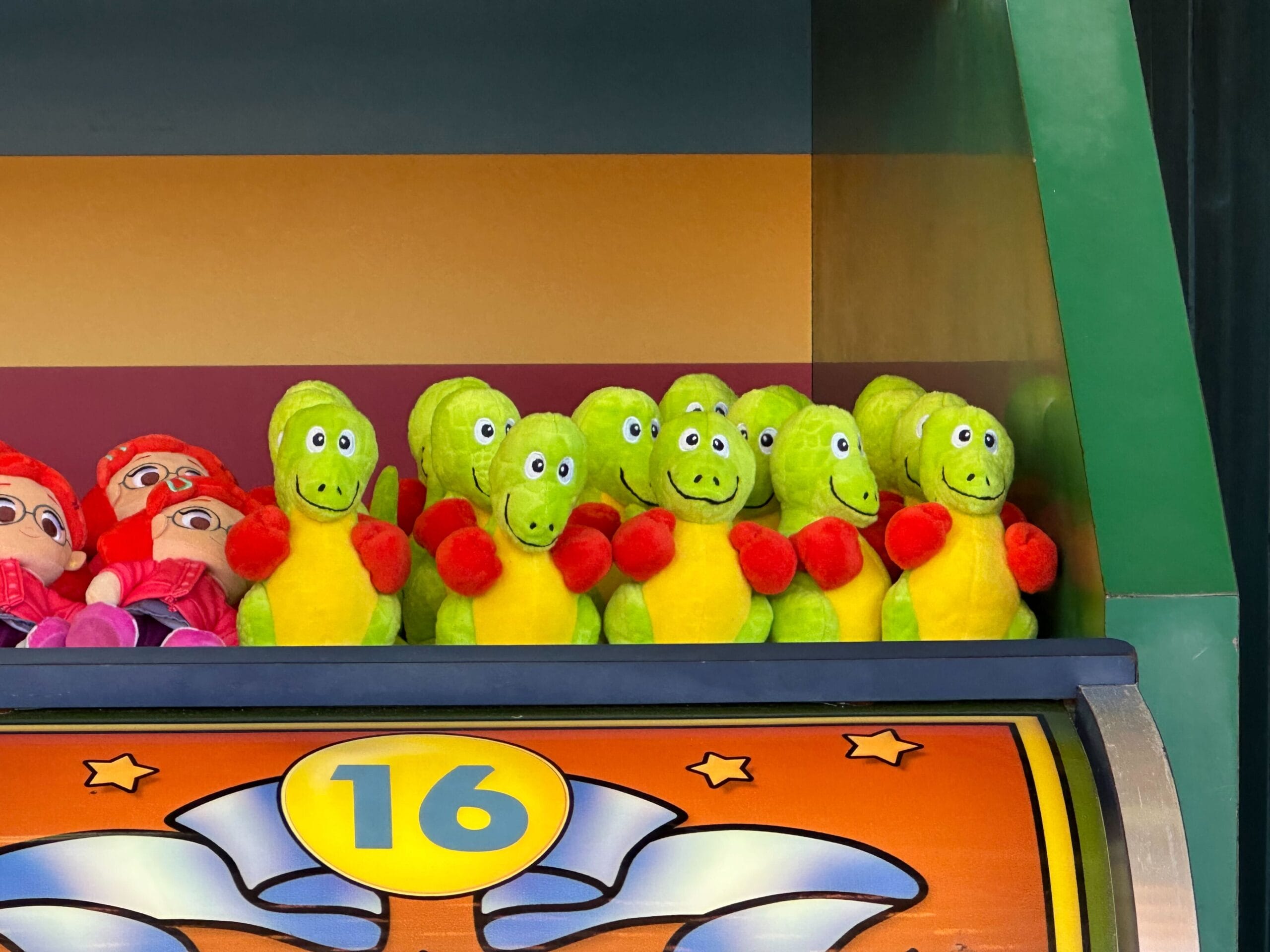 A row of green dinosaur plush toys with red arms is displayed on a carnival game shelf in DinoLand, labeled with the number 16.