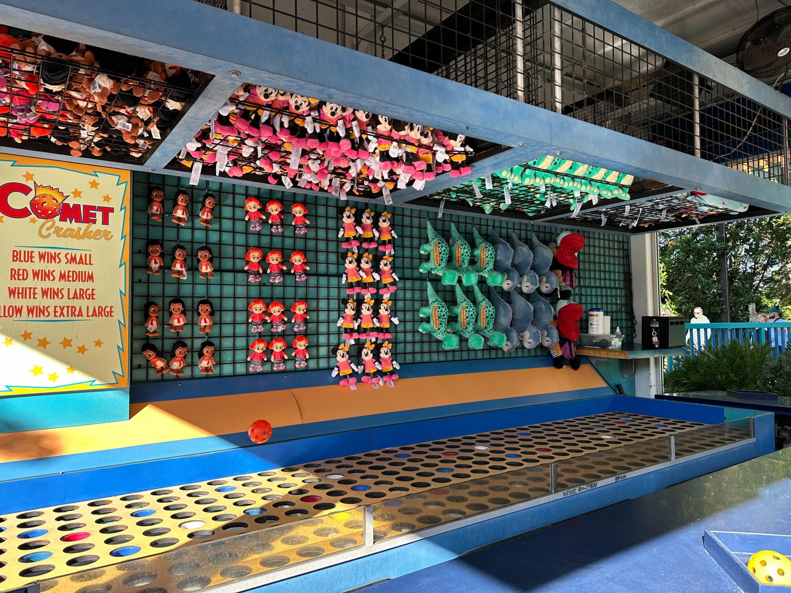 Fossil Fun Games booth in DinoLand features walls of plush toys, a prize sign on the left, and circular targets on the counter. Sunlight illuminates the scene.
