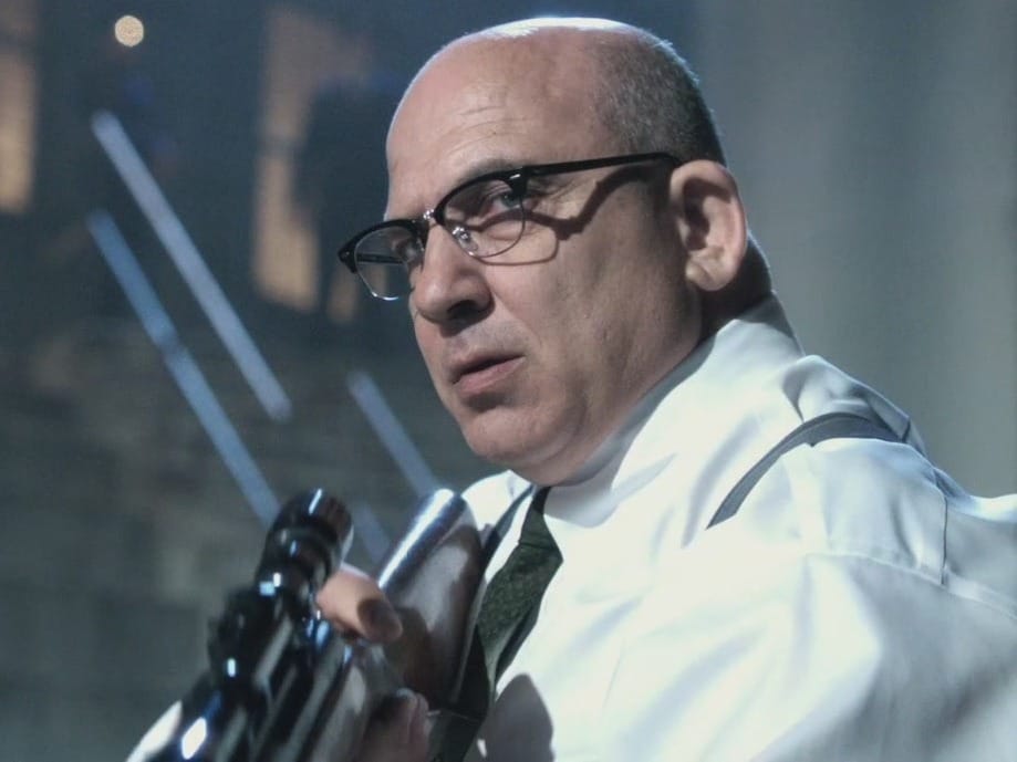 A bald man wearing glasses, resembling a Mandalorian warrior, is holding a large metallic object on his shoulder, looking off to the side.