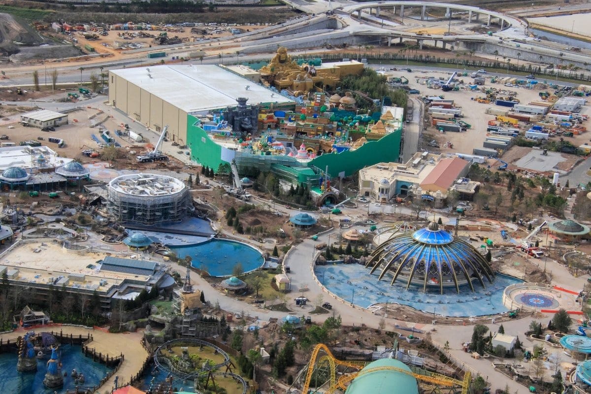 Aerial view of Epic Universe theme park under construction, featuring large buildings, roller coasters, and various themed attractions surrounded by roads and construction vehicles.