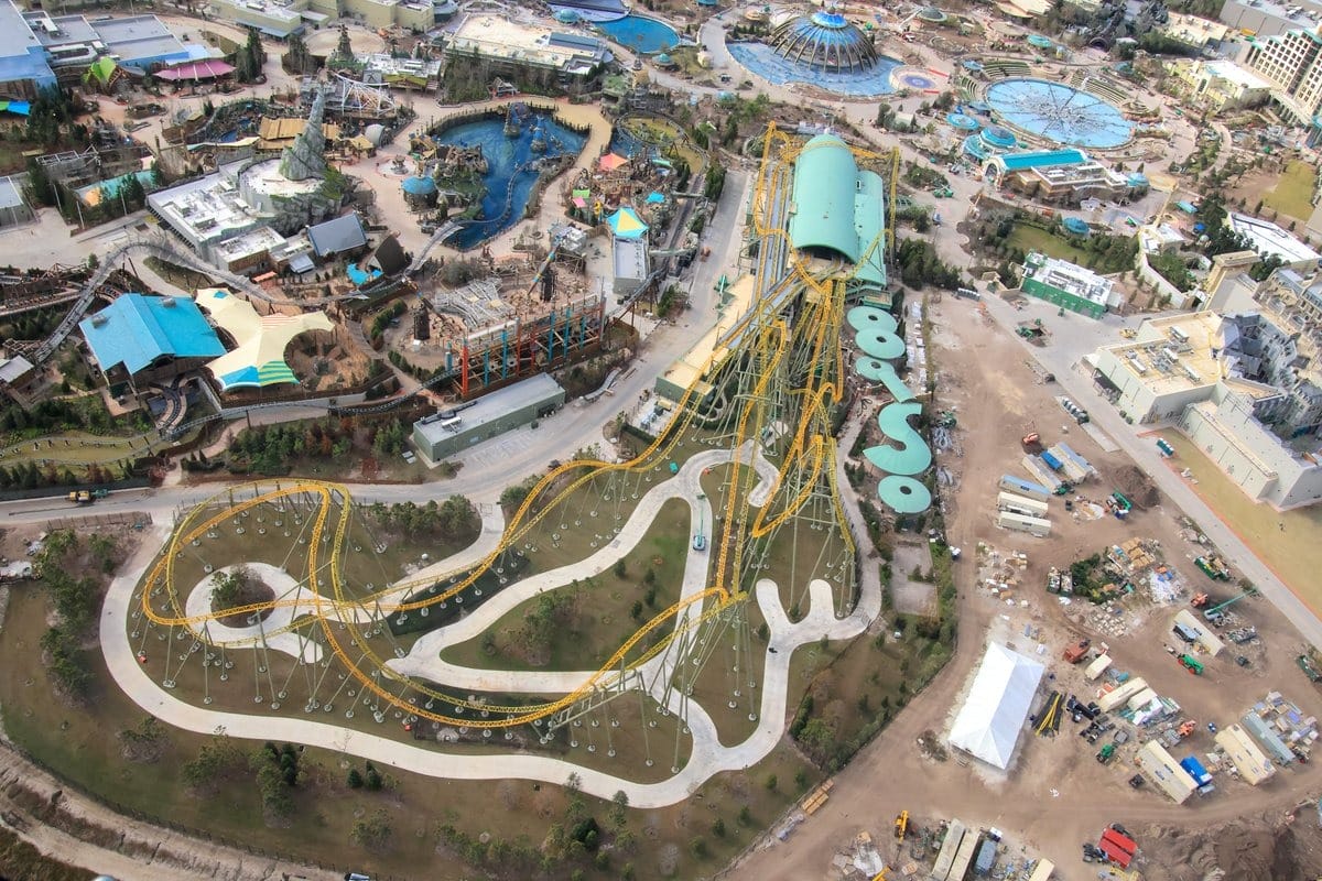 Aerial view of an epic theme park featuring a large roller coaster with yellow tracks, various buildings, pathways, and landscaped areas.