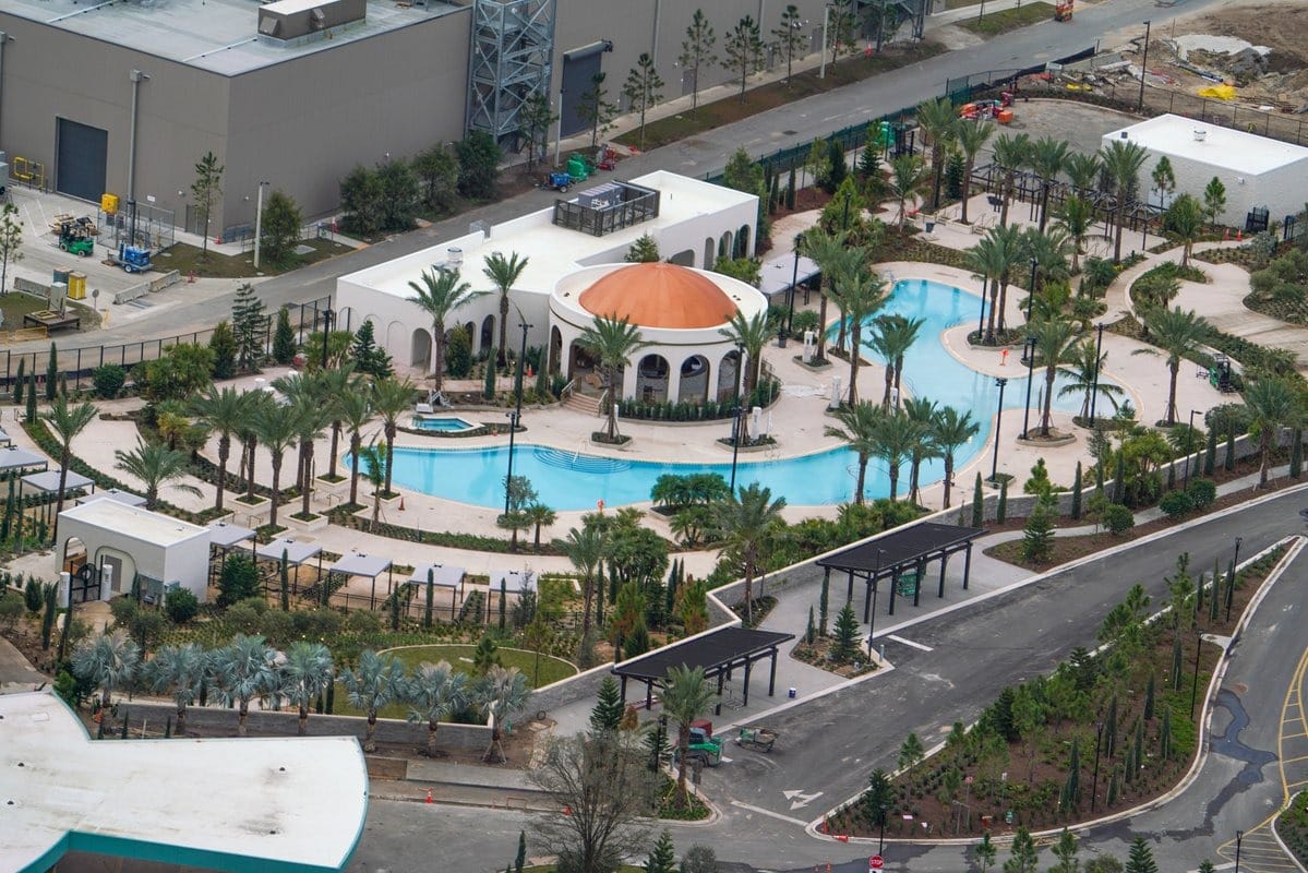 From an aerial view, the landscaped area reveals a winding blue pool and palm trees, seamlessly blending with pathways amongst surrounding buildings. In the distance, hints of Epic Universe come into sight, adding a touch of magic to this picturesque scene.