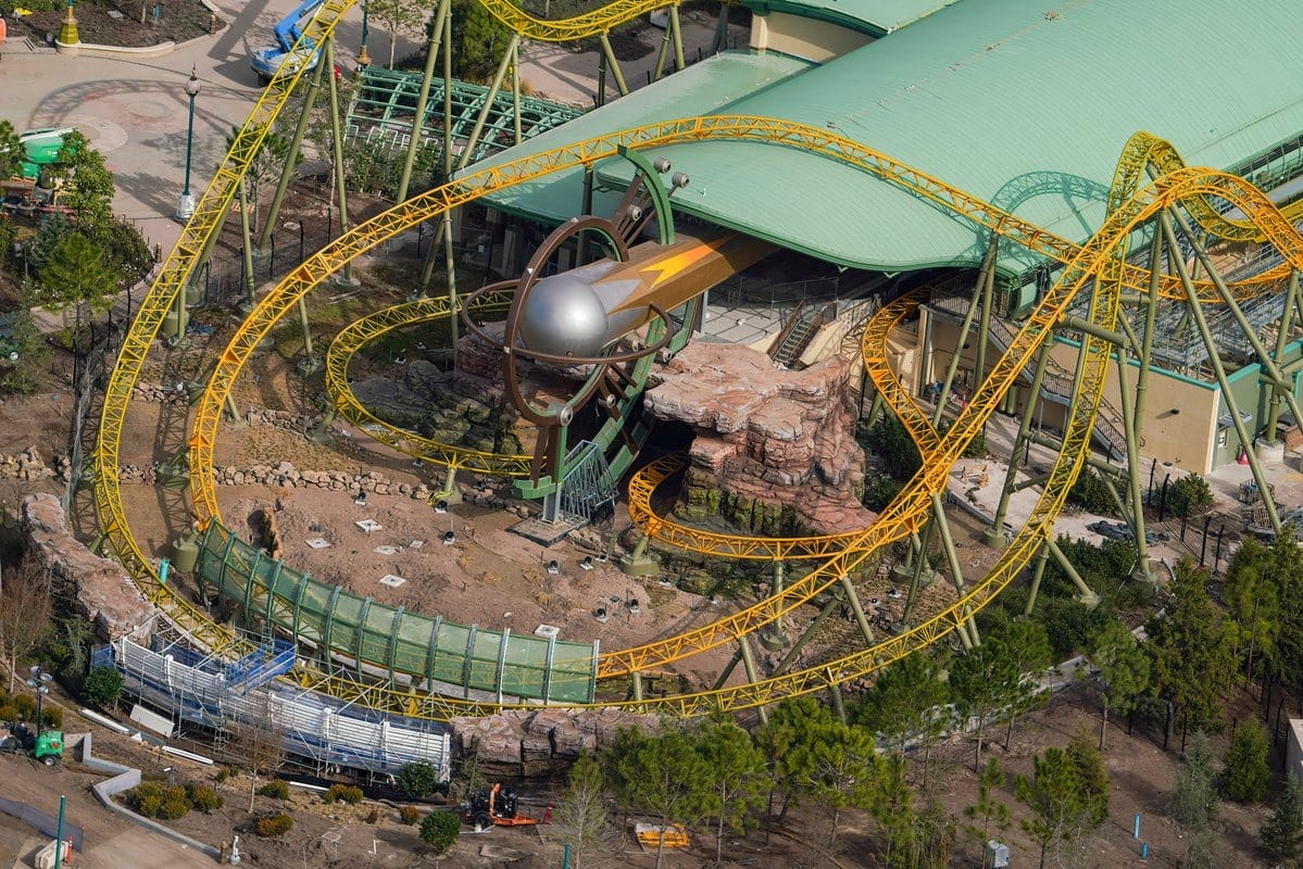 Aerial view of an Epic Universe theme park features a roller coaster with yellow tracks and a metallic rocket-themed design, surrounded by trees and structures.