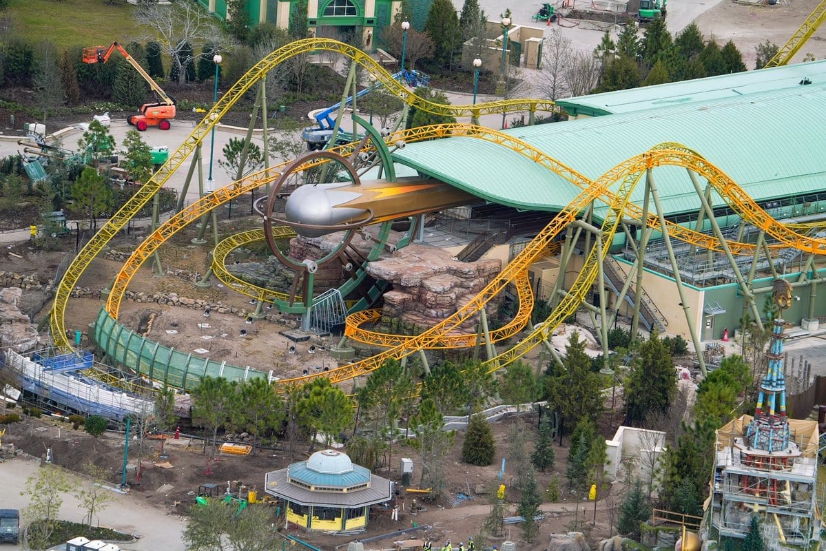 Aerial view of Epic Universe's new theme park ride under construction, featuring winding yellow tracks and a large building with a green roof. Construction equipment and workers are visible around the site.