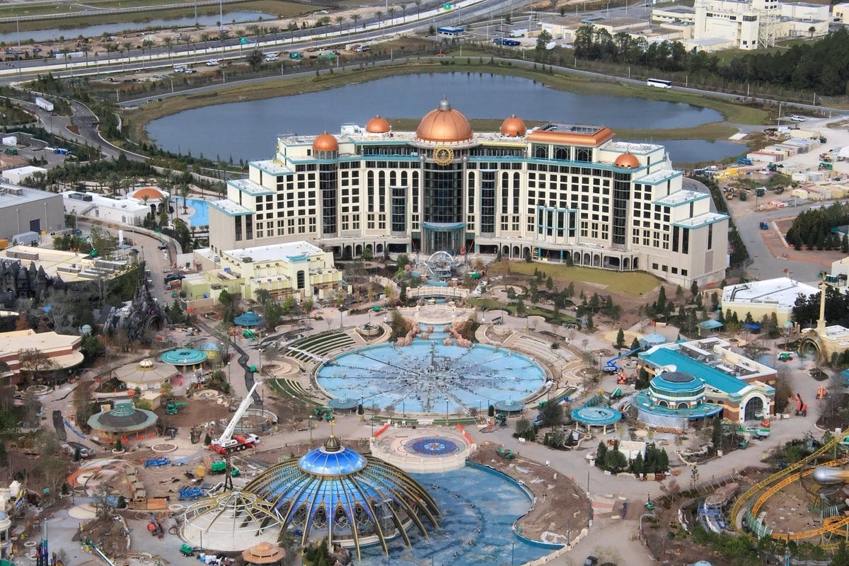 An aerial view reveals a large, domed hotel amidst landscaped grounds and water features, with construction equipment visible in the foreground, hinting at the nearby Epic Universe theme park.
