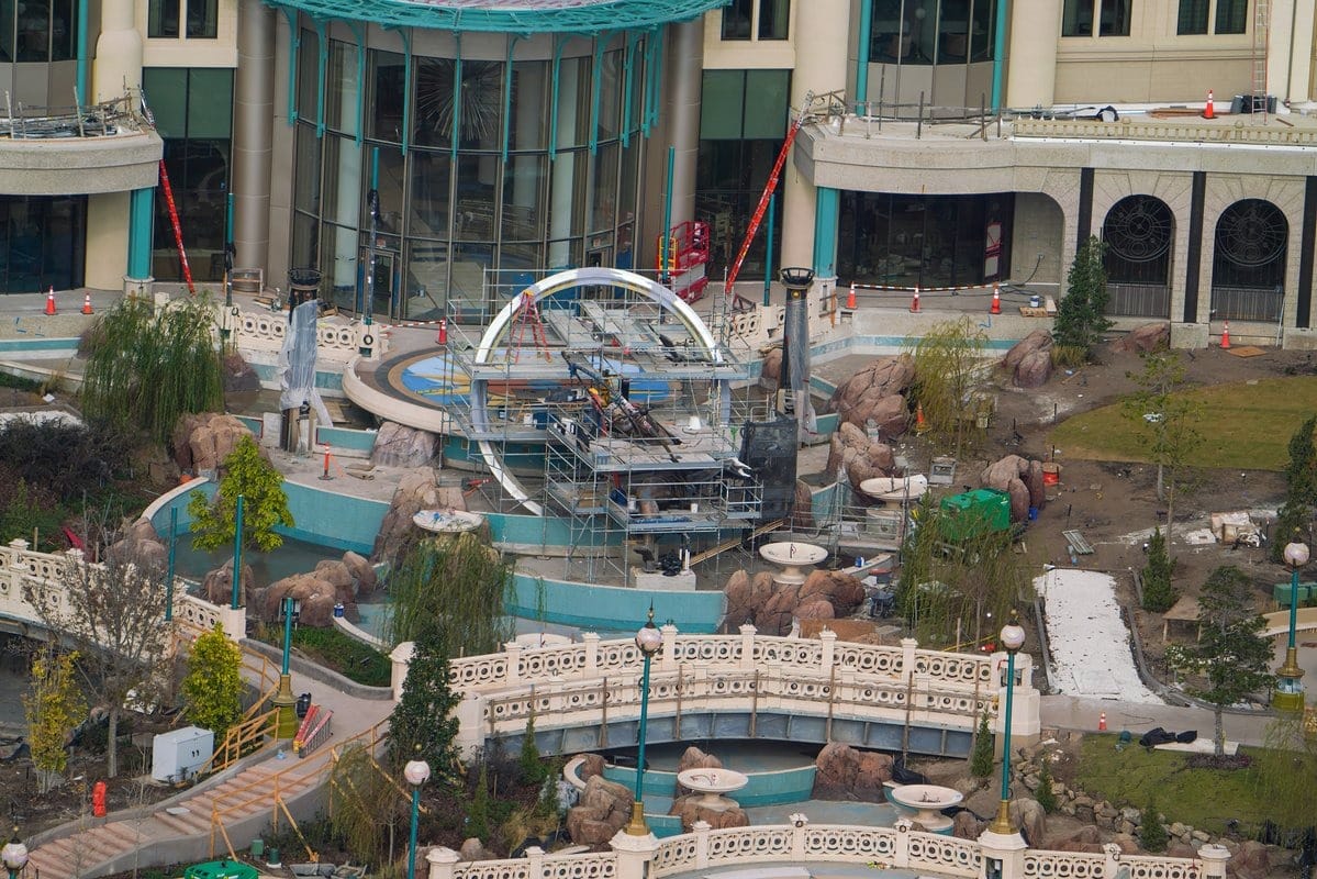 The construction site of a large building features an epic universe theme with a fountain centerpiece, scaffolding, and landscaping. Pathways twist through lush greenery, creating a mesmerizing blend of nature and architecture.
