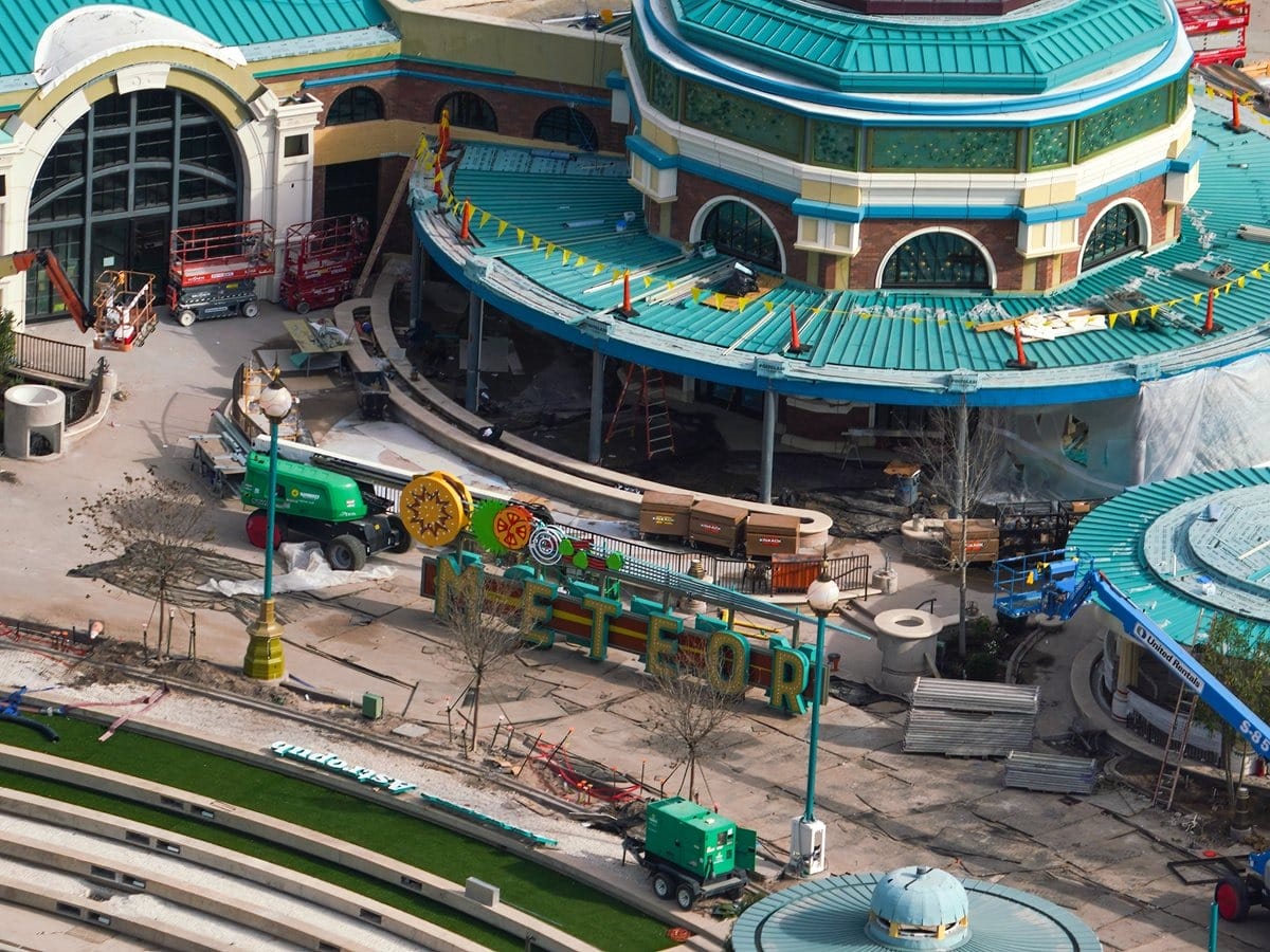 Aerial view of a construction site with people, vehicles, and colorful signs spelling "METEO" in front of a circular building with a turquoise roof, all set against the backdrop of an epic universe unfolding beyond.