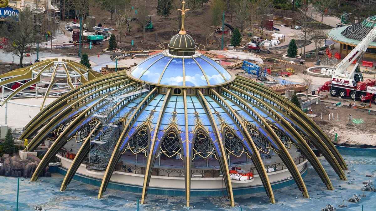 Aerial view of a large, glass-domed building under construction, reminiscent of an epic universe. It features golden rib-like supports and is surrounded by construction equipment and materials.