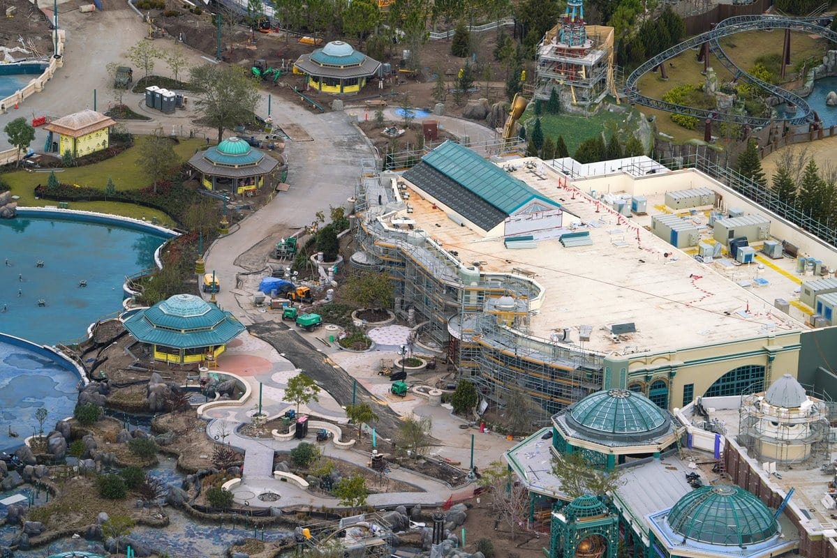 Aerial view of an epic universe theme park under construction with various buildings, pathways, water features, and landscaping in progress. Scaffolding and construction equipment are visible.